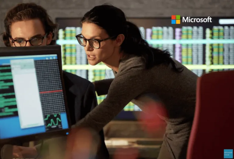 Two people wearing glasses look at a computer screen displaying data and graphs; a woman leaning forward points at the screen while a man watches. The Microsoft logo appears in the top right corner.