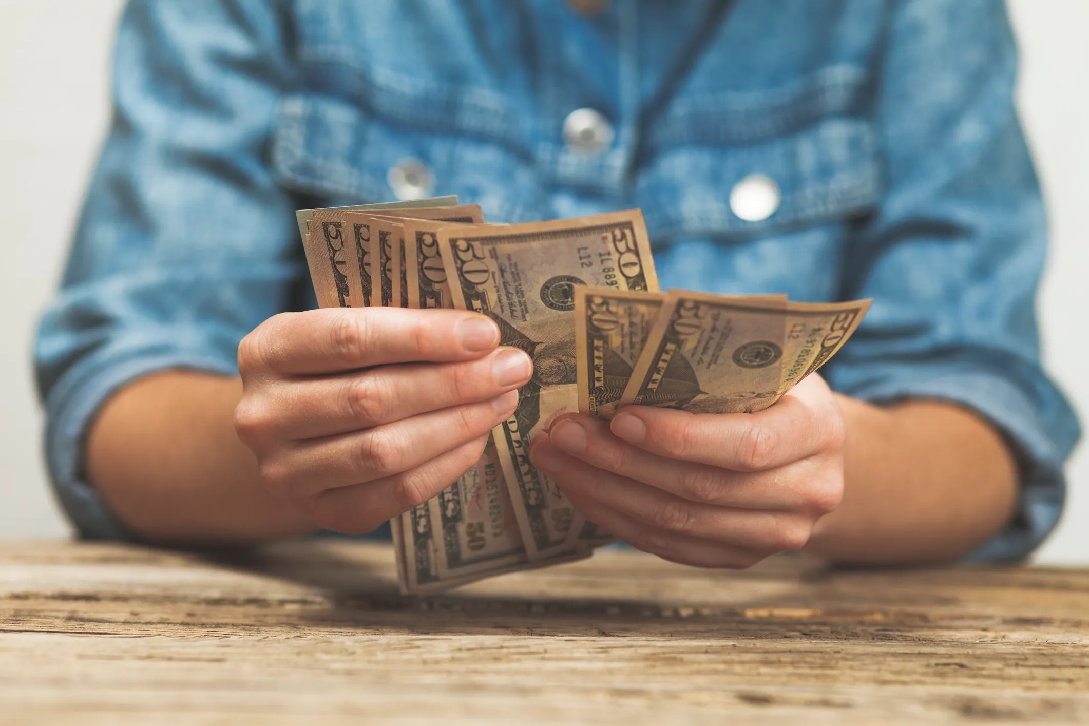 Person wearing a blue denim shirt counting multiple 50-dollar bills over a wooden table.