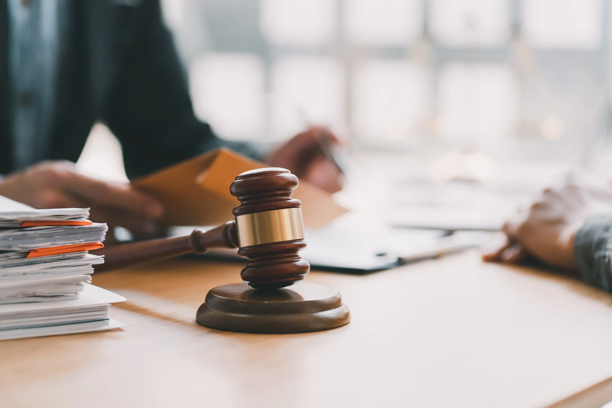 Close-up of a wooden judge's gavel on a desk beside a stack of documents, with two people in the blurred background reviewing papers.
