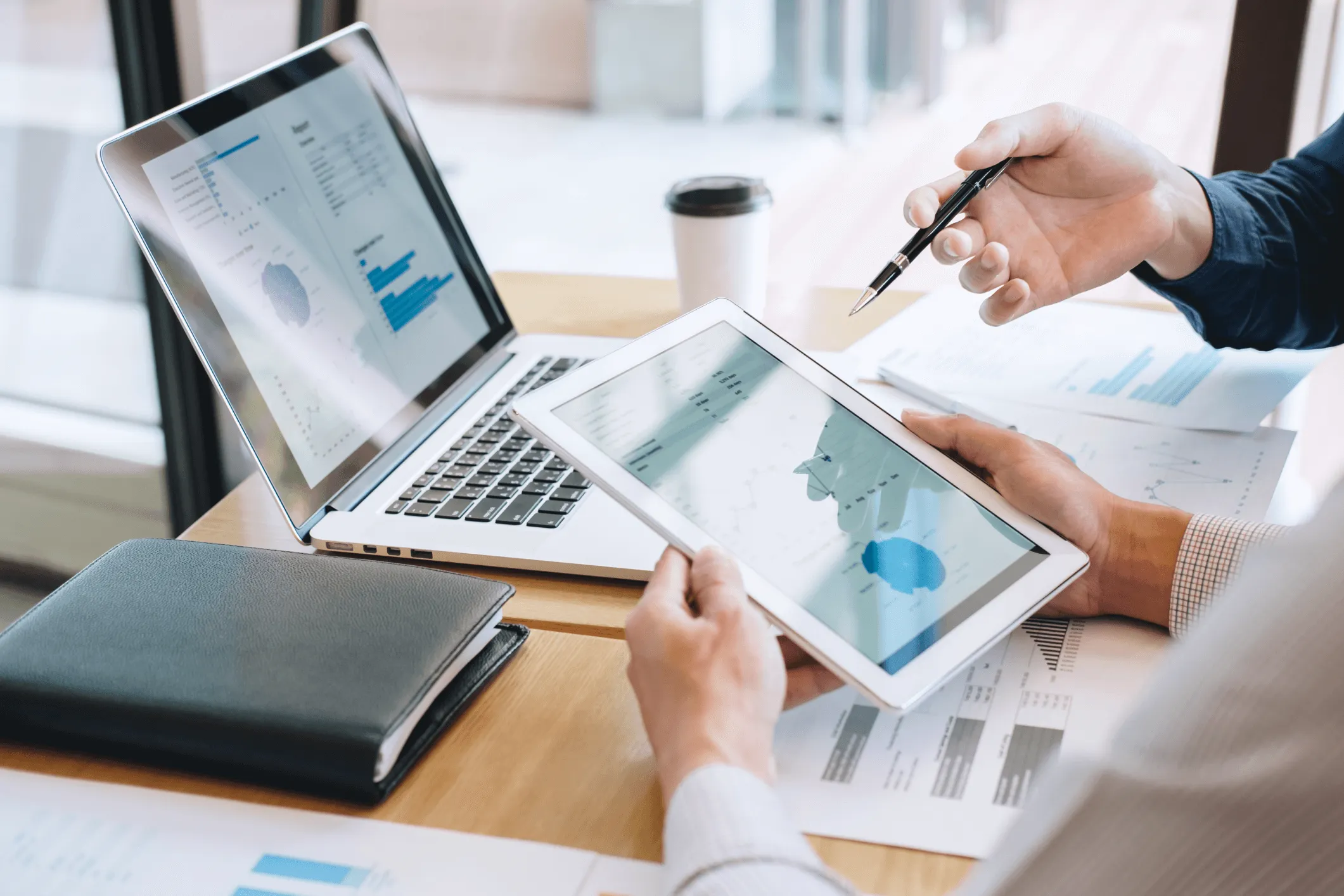 Two people reviewing financial data displayed on a tablet and a laptop, with printed charts and a notebook on a wooden table, and a coffee cup in the background.