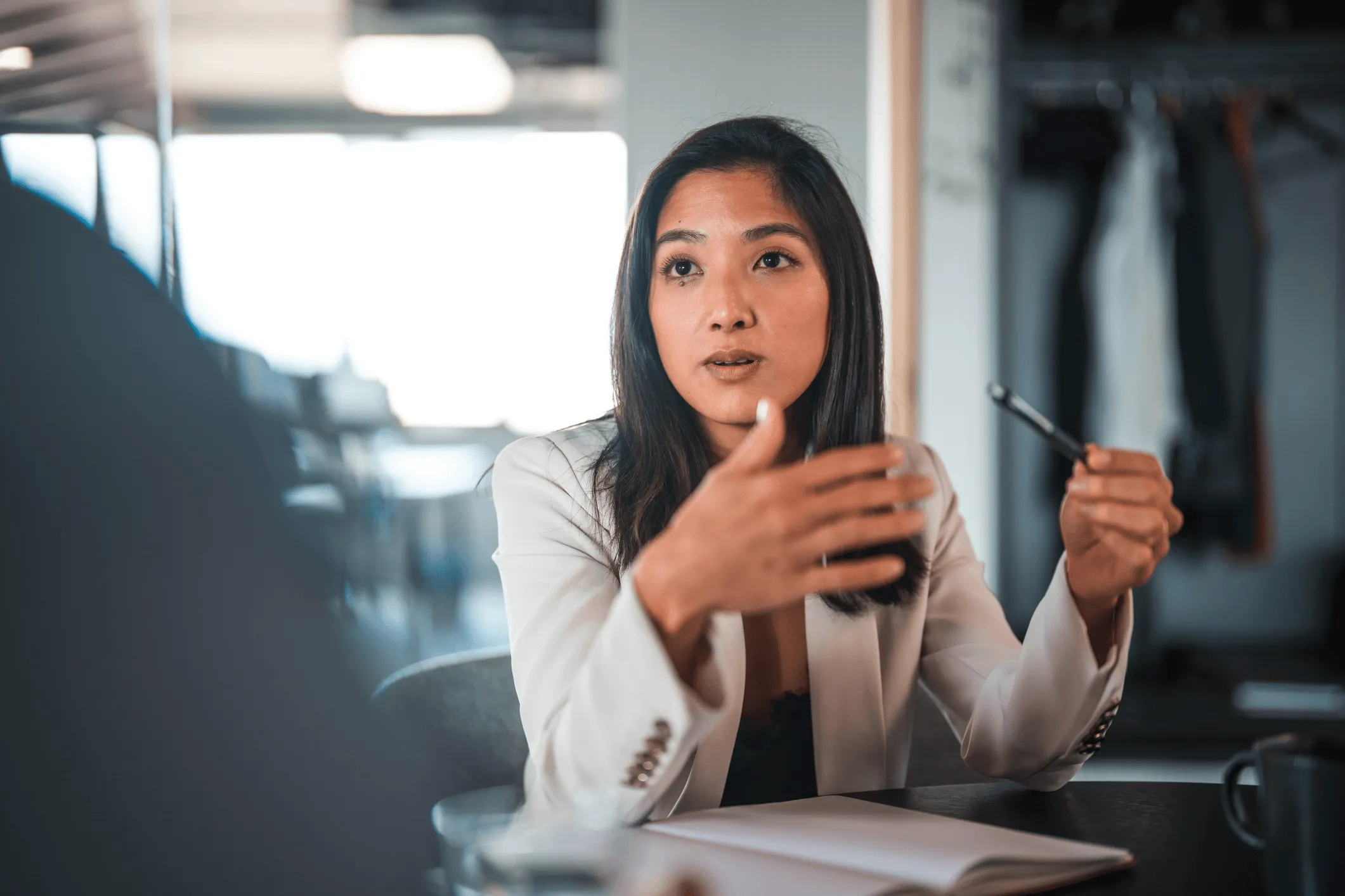 Woman in a white blazer speaking and gesturing with a pen during a discussion in an office setting.