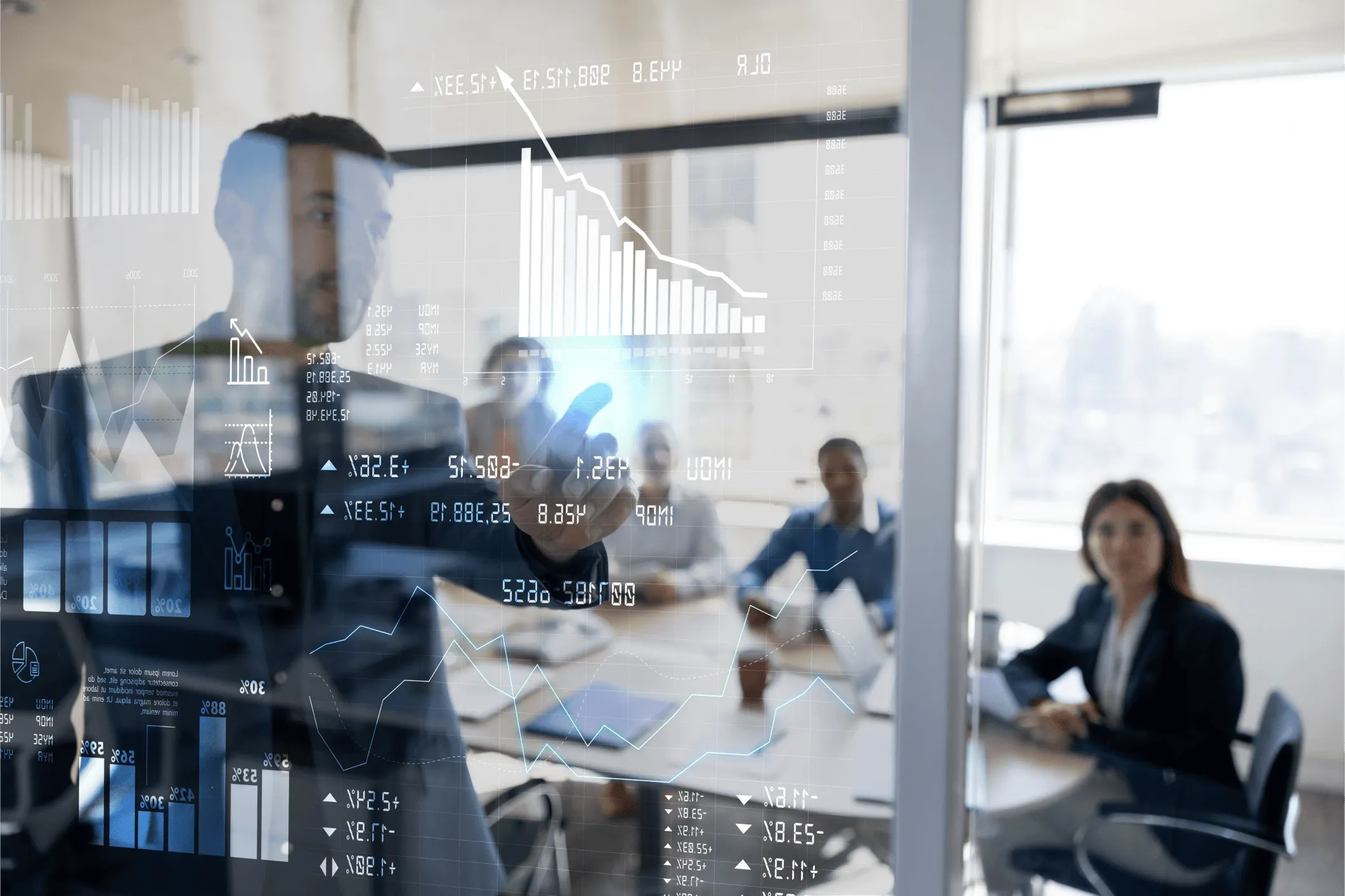 Businessman in an office pointing at a transparent screen displaying digital financial charts and data, with a group of professionals seated around a conference table in the background.