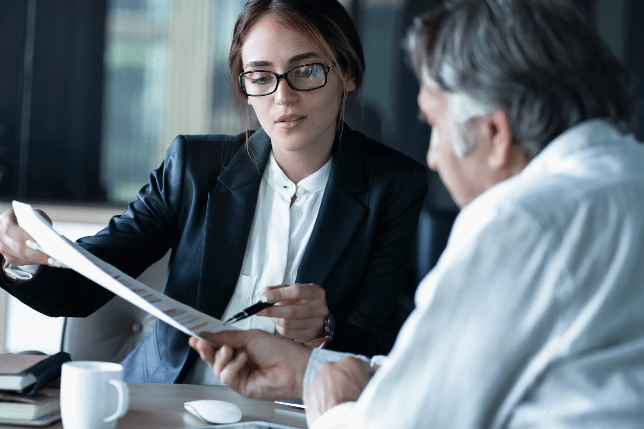 A businesswoman wearing glasses and a black blazer pointing at a document with a pen while discussing it with an older man in a white shirt at a desk with a coffee cup and books.