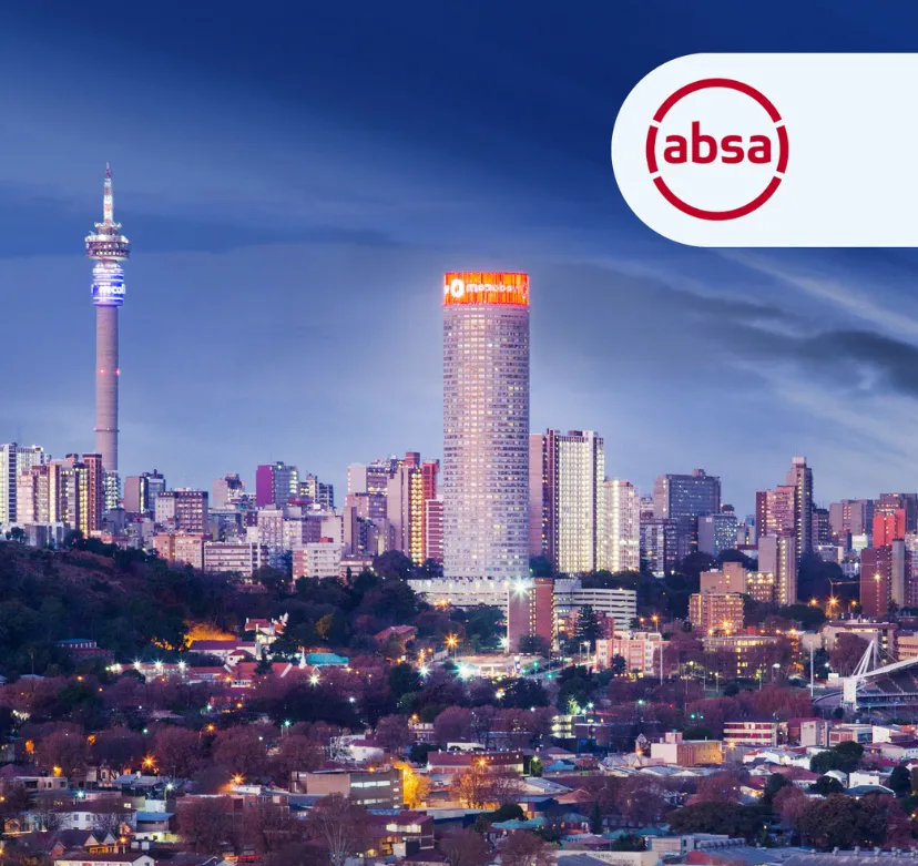 Cityscape at dusk with a tall communication tower on the left and a cylindrical skyscraper with illuminated signage near the center, surrounded by many buildings and trees under a clear sky.