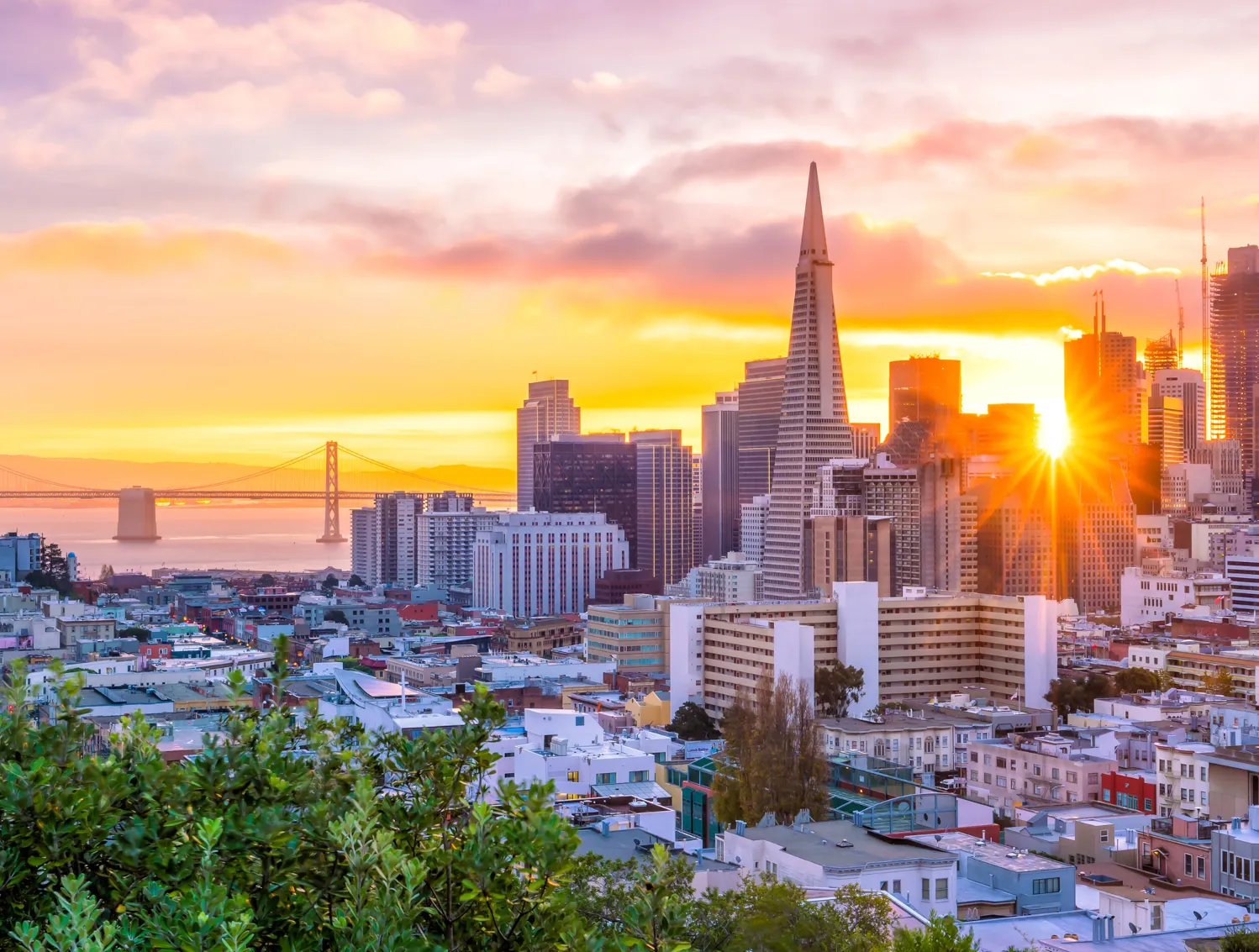 San Francisco cityscape at sunset with the Transamerica Pyramid prominently in the skyline and the Bay Bridge in the background, with sunlight casting a warm glow over the buildings.