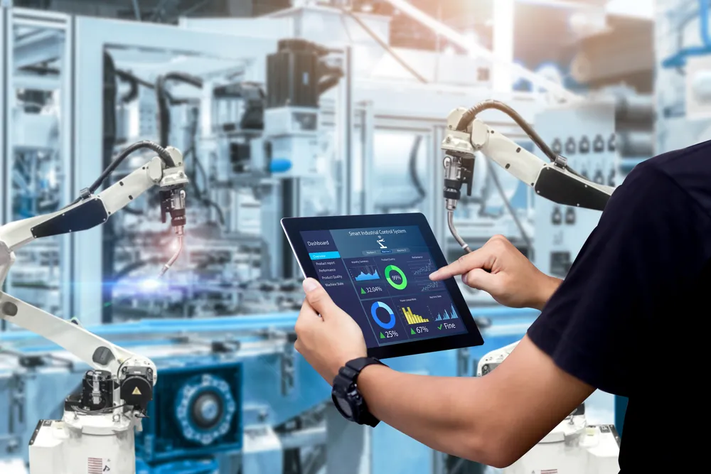 Person holding a tablet displaying a smart industrial control system dashboard with graphs and charts, in front of robotic arms operating in a high-tech factory setting.