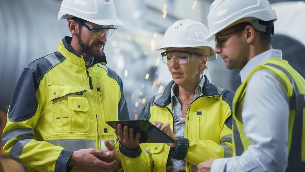 Three construction workers wearing hard hats, safety glasses, and high-visibility jackets reviewing information on a digital tablet indoors with sparks flying in the background.