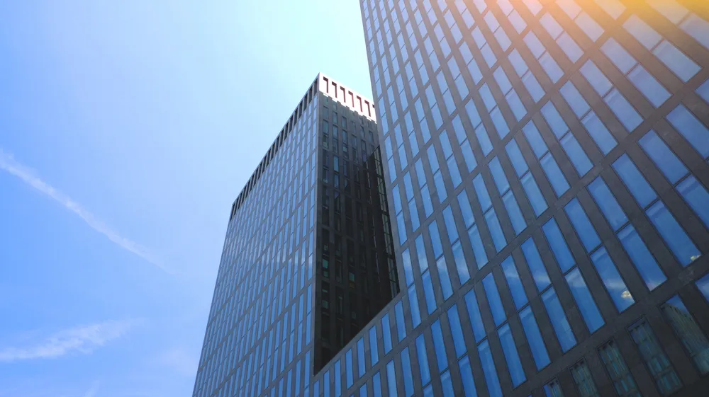 Low-angle view of two modern glass skyscrapers reflecting the clear blue sky with sunlight in the upper right corner.