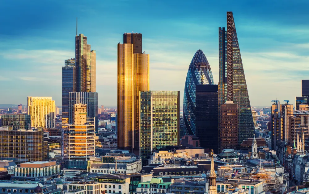 Cityscape of London featuring iconic skyscrapers including the Gherkin, the Leadenhall Building, and other modern high-rises under a partly cloudy blue sky during golden hour.