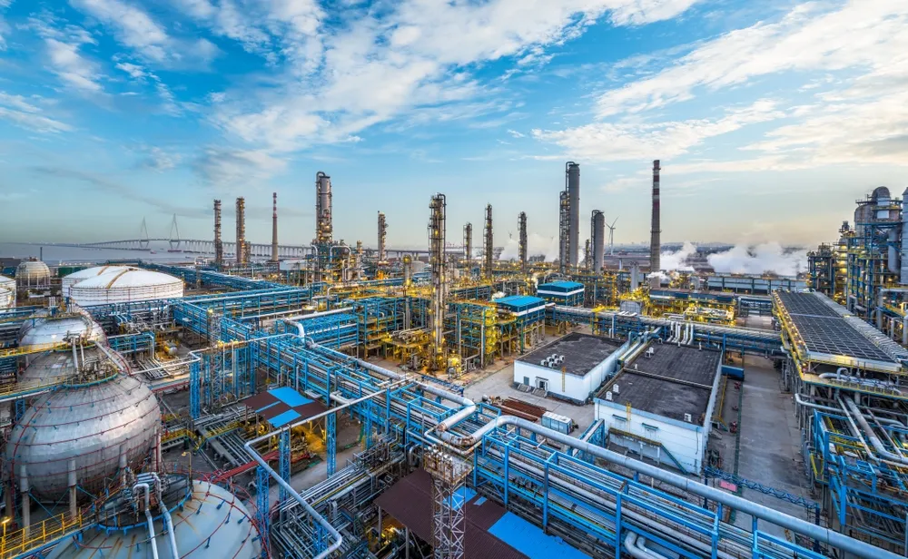Panoramic view of a large industrial oil refinery complex with multiple pipelines, storage tanks, tall distillation towers, and smokestacks releasing steam under a partly cloudy blue sky near a waterfront.