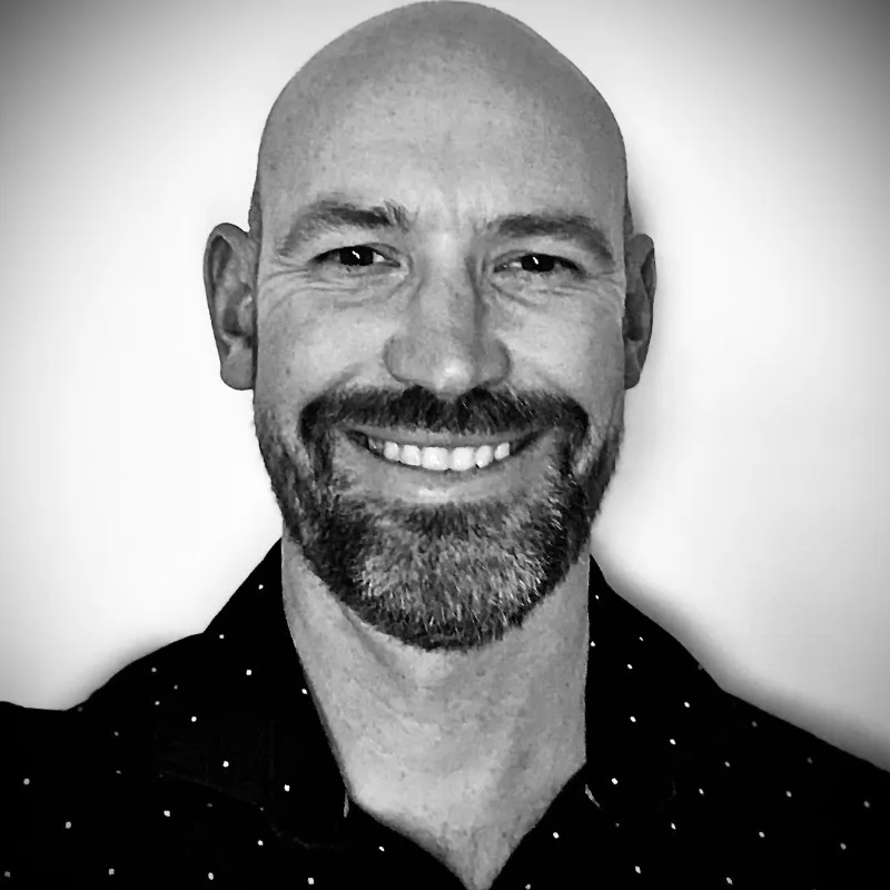 Black and white close-up portrait of a bald man with a beard and mustache smiling at the camera, wearing a dark shirt with small white polka dots.
