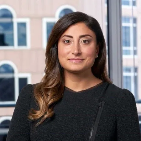 Professional woman with long, wavy brown hair wearing a black top, standing in front of a window showing a building exterior.