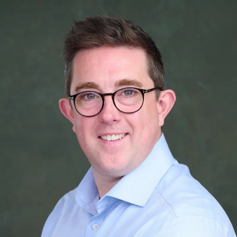 Smiling man with short brown hair and glasses wearing a light blue collared shirt against a dark green background.