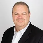 Smiling middle-aged man with short hair wearing a white shirt and black blazer, against a plain light gray background.