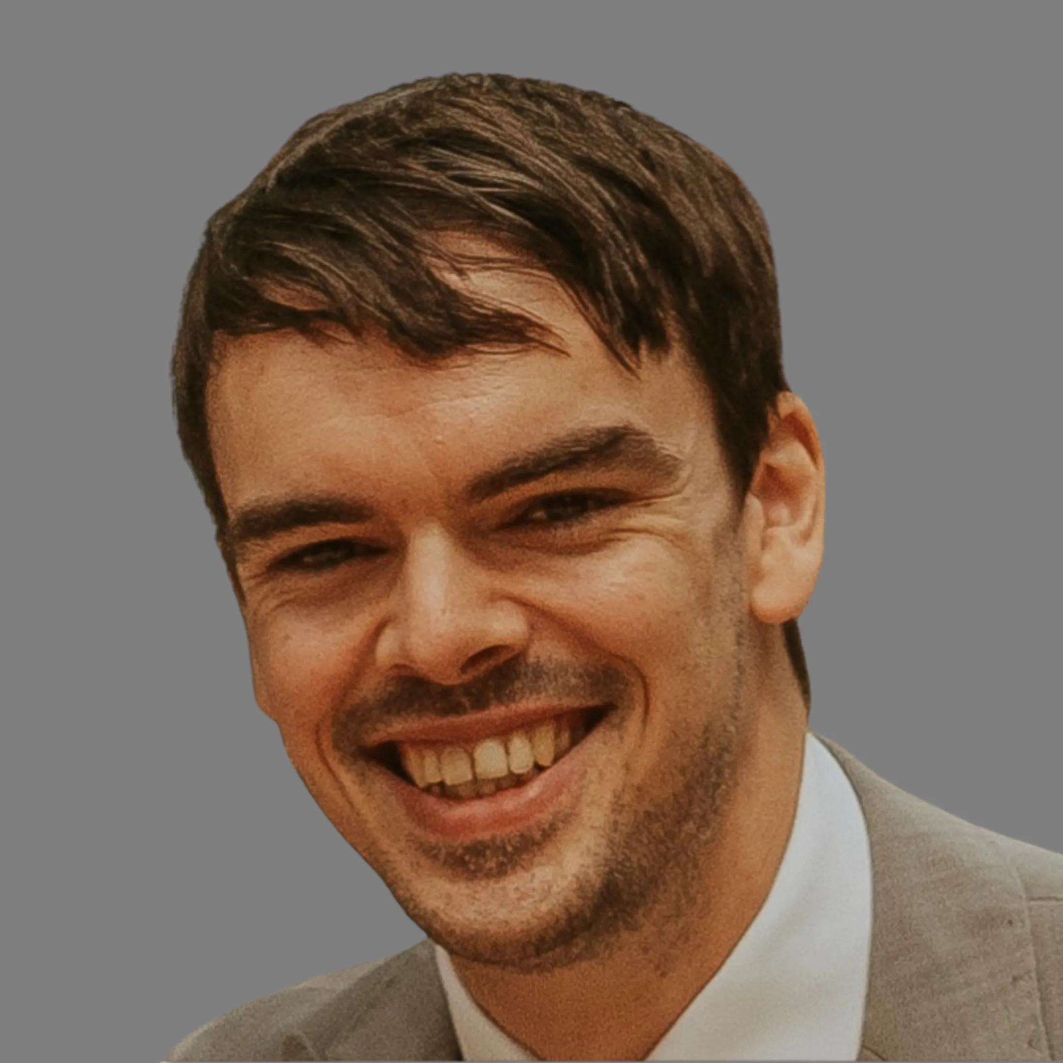 Close-up of a smiling man with short dark hair and light facial hair wearing a light-colored suit jacket and white shirt against a gray background.