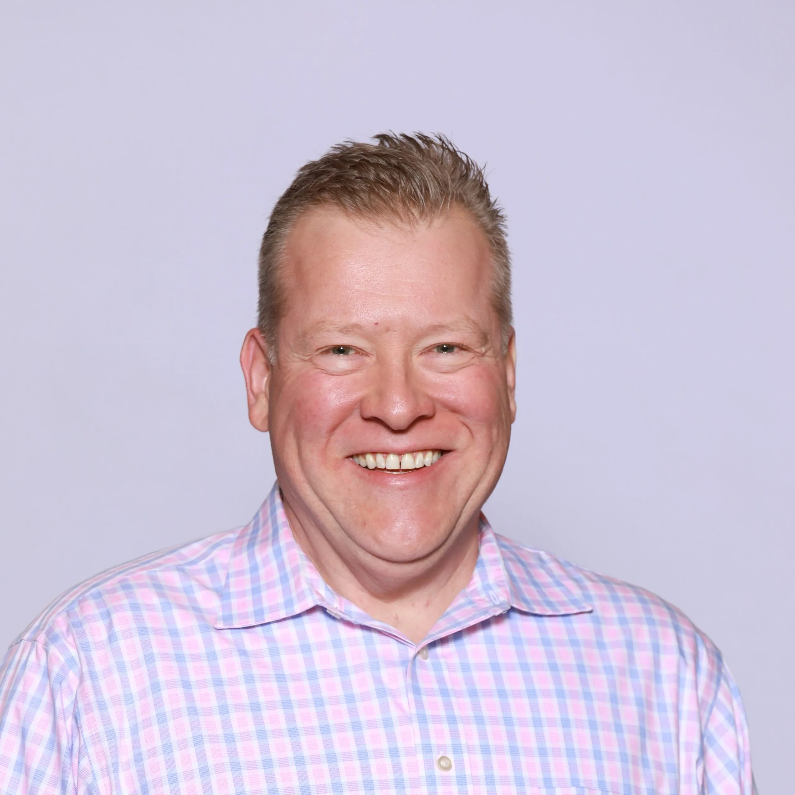 Smiling middle-aged man with short light brown hair wearing a pink and blue checkered button-up shirt against a plain light purple background.