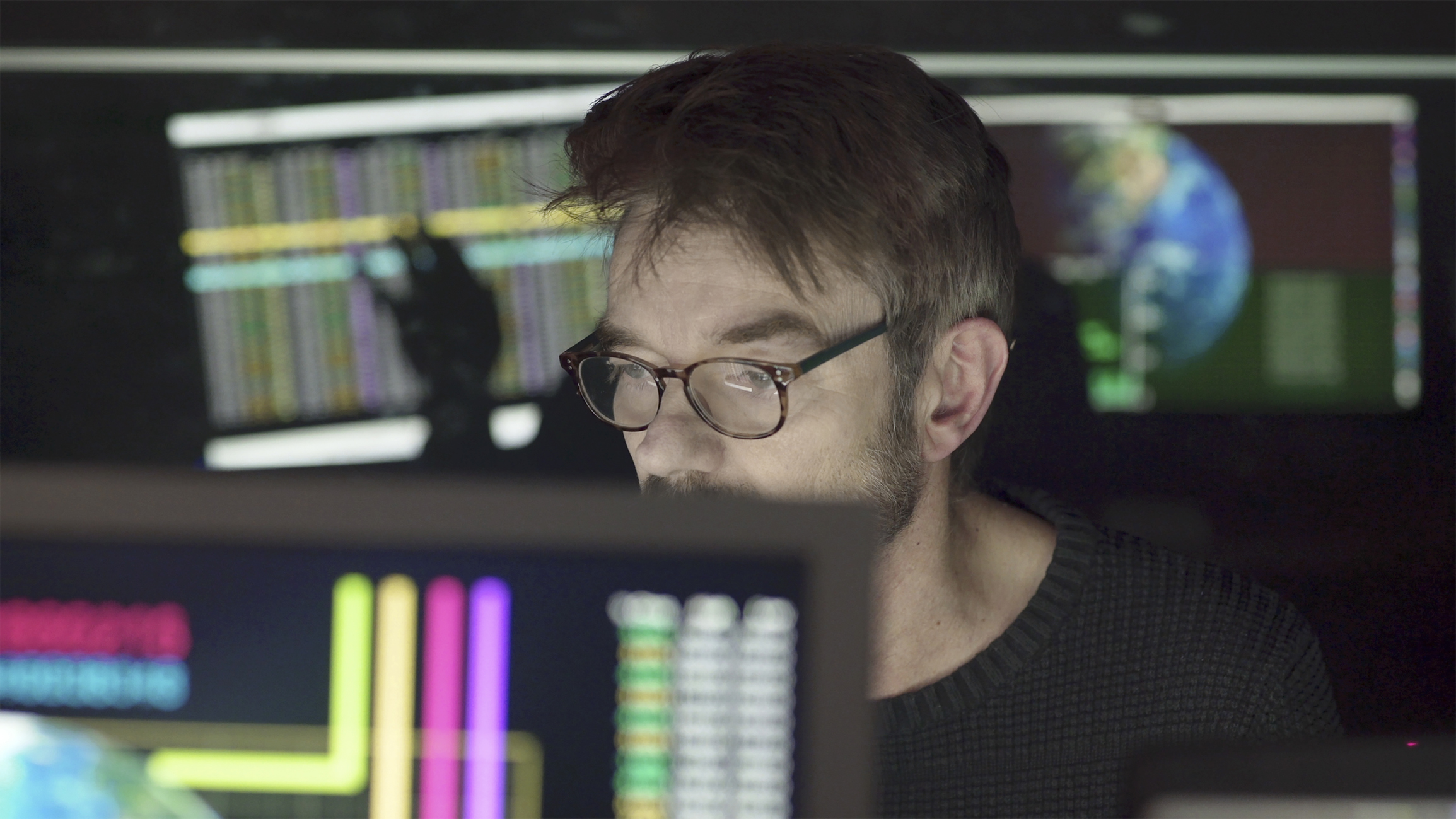 Man wearing glasses intently looks at multiple computer screens displaying colorful data charts and a globe, in a dark room.