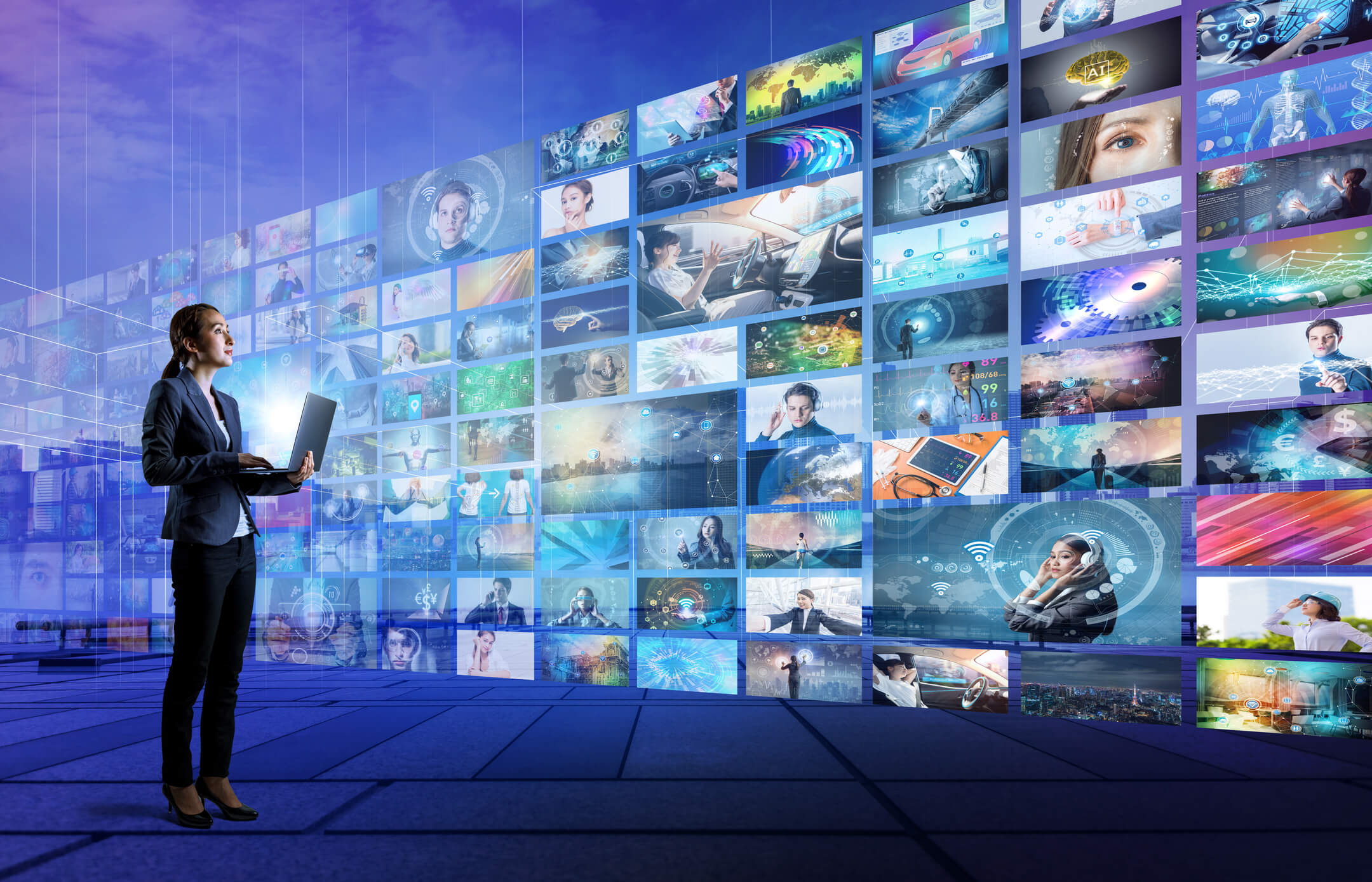 A businesswoman holding a laptop stands in a digital room with a large wall of multiple futuristic and tech-themed images, including AI, medical, automotive, and data visuals.