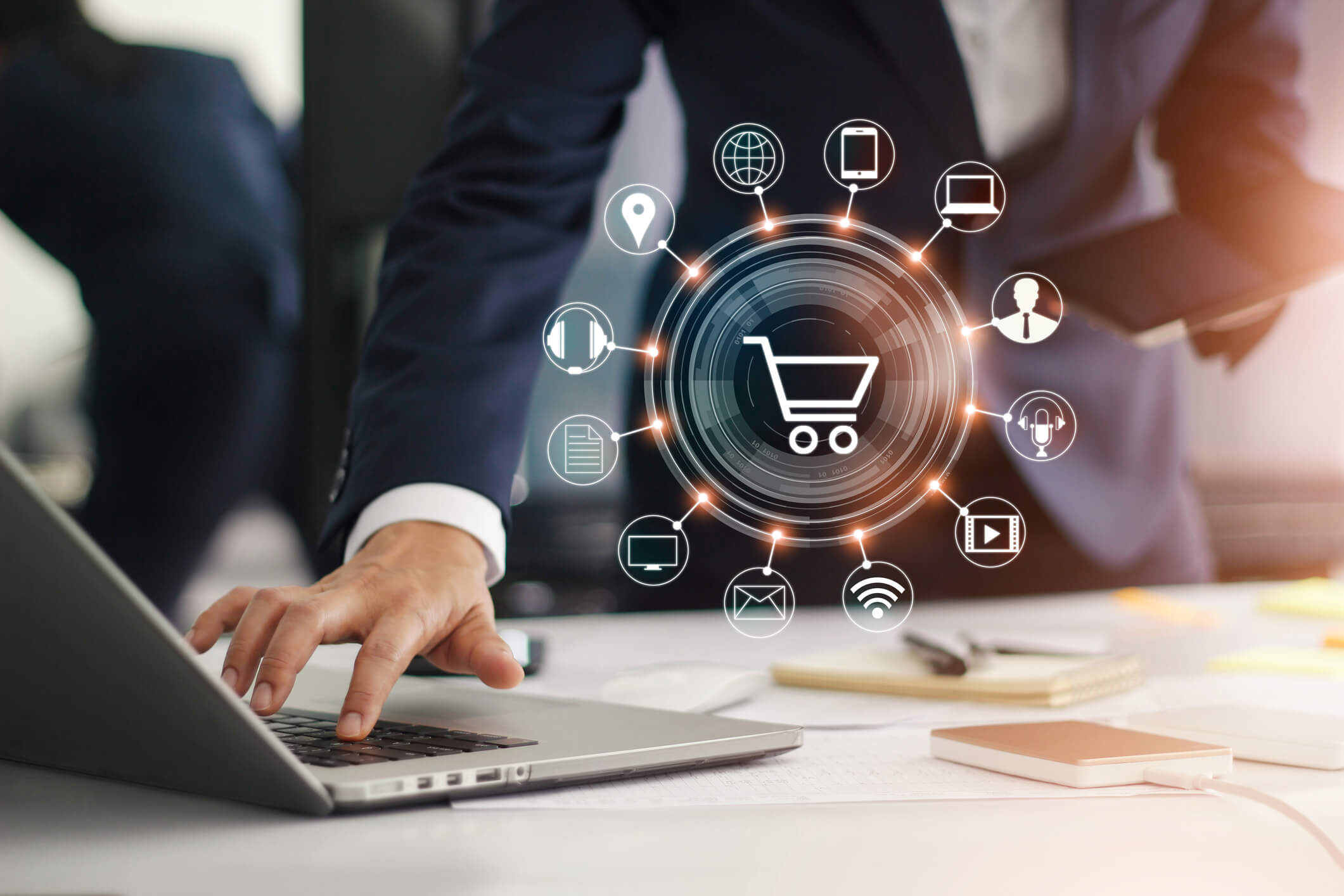Person in business attire using a laptop on a desk with a holographic shopping cart icon surrounded by technology and communication symbols like globe, mobile phone, laptop, and wifi, representing online retail or e-commerce.