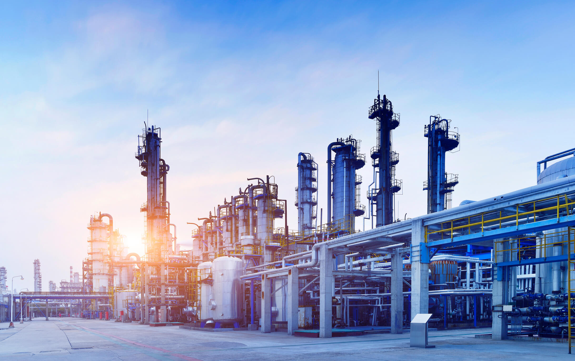 Industrial chemical plant with tall distillation towers, pipelines, and storage tanks under a clear blue sky at sunrise or sunset.