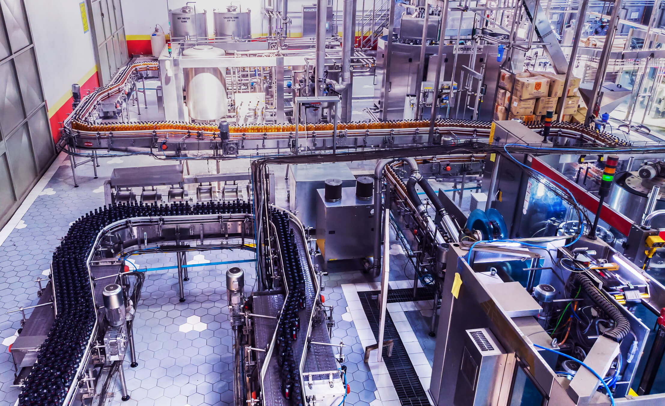 High-angle view of an automated beverage bottling factory with conveyor belts carrying dark and orange bottles through stainless steel machinery and equipment on a hexagonal tiled floor.