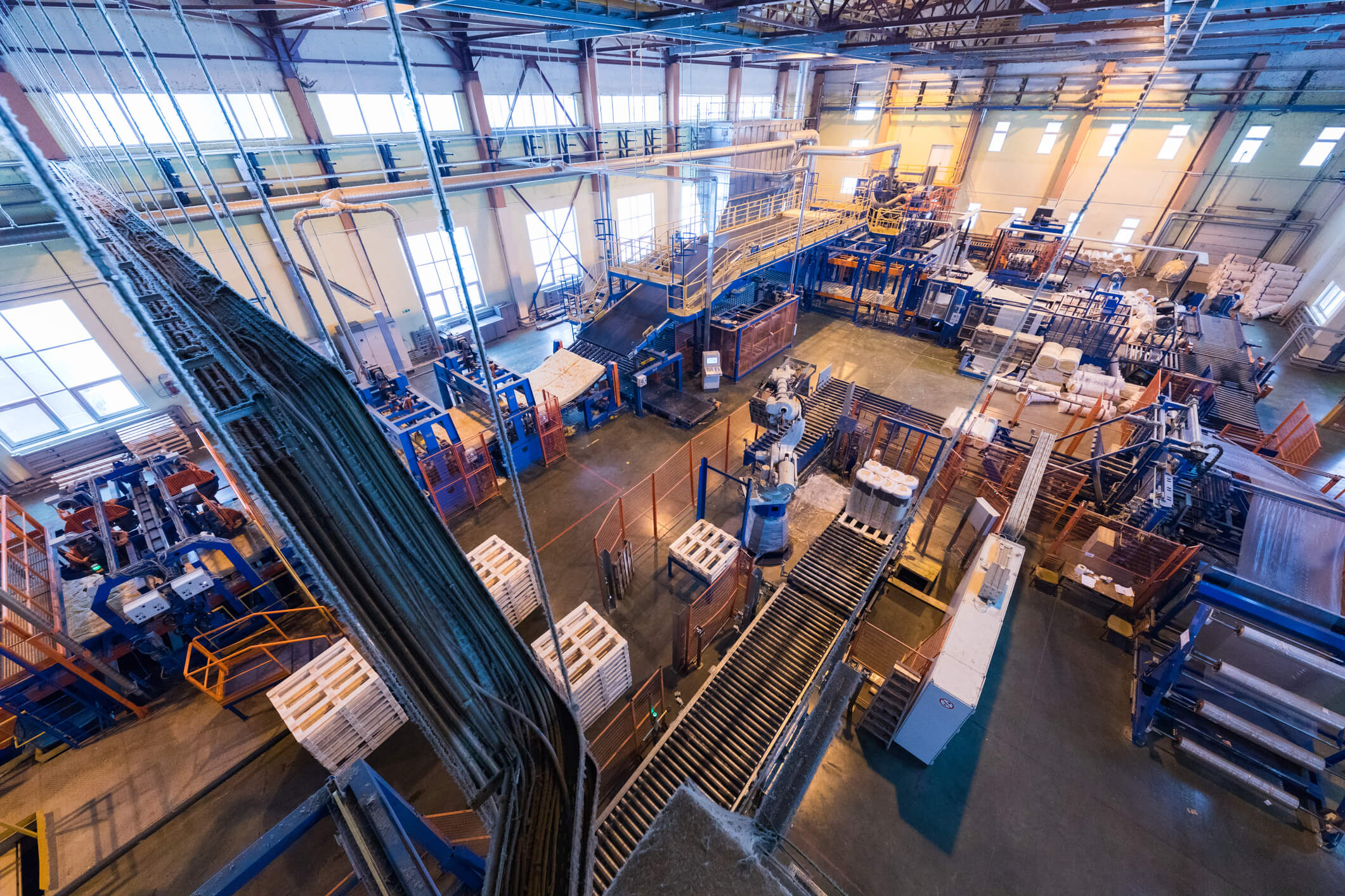 Interior view of a large industrial manufacturing facility with automated conveyor belts, robotic arms, and stacks of materials, illuminated by natural and artificial light.