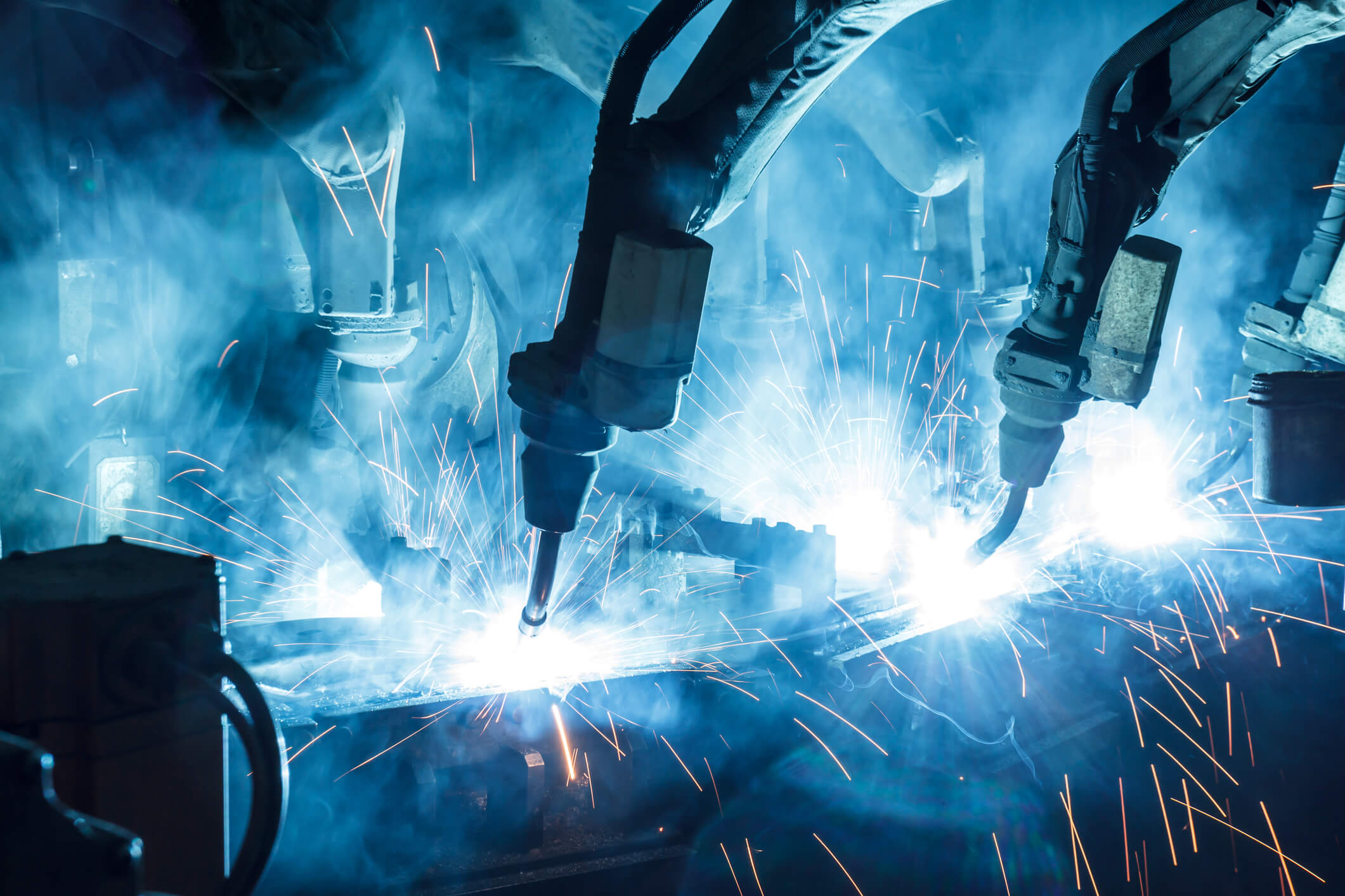 Close-up of industrial robotic arms performing welding, emitting bright sparks and smoke in a factory setting.