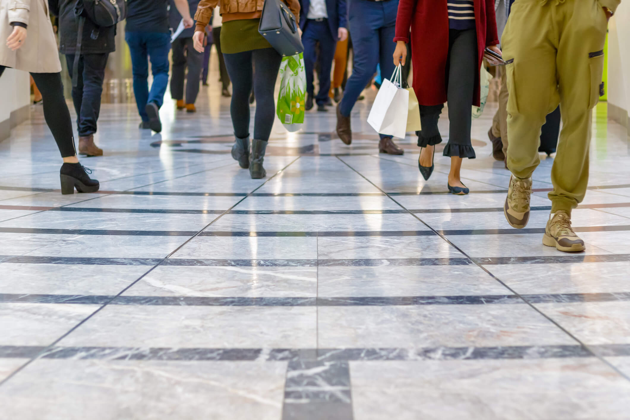 People walking in a shopping mall corridor with shiny tiled floors; two individuals are carrying shopping bags.