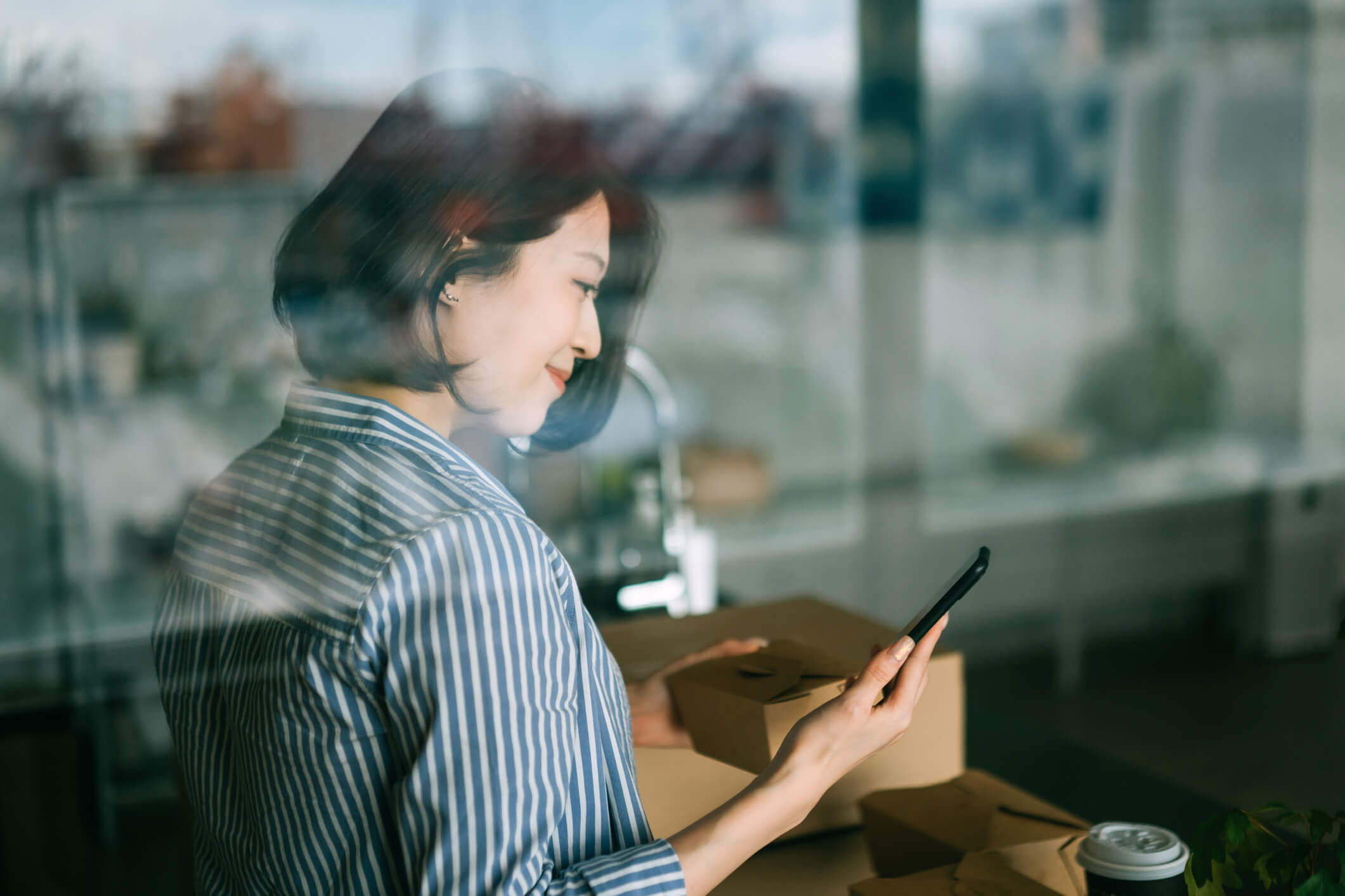 Smiling woman wearing a blue and white striped shirt inside a room, holding a smartphone in one hand and a small cardboard box in the other, with more cardboard boxes and a coffee cup on a table nearby.