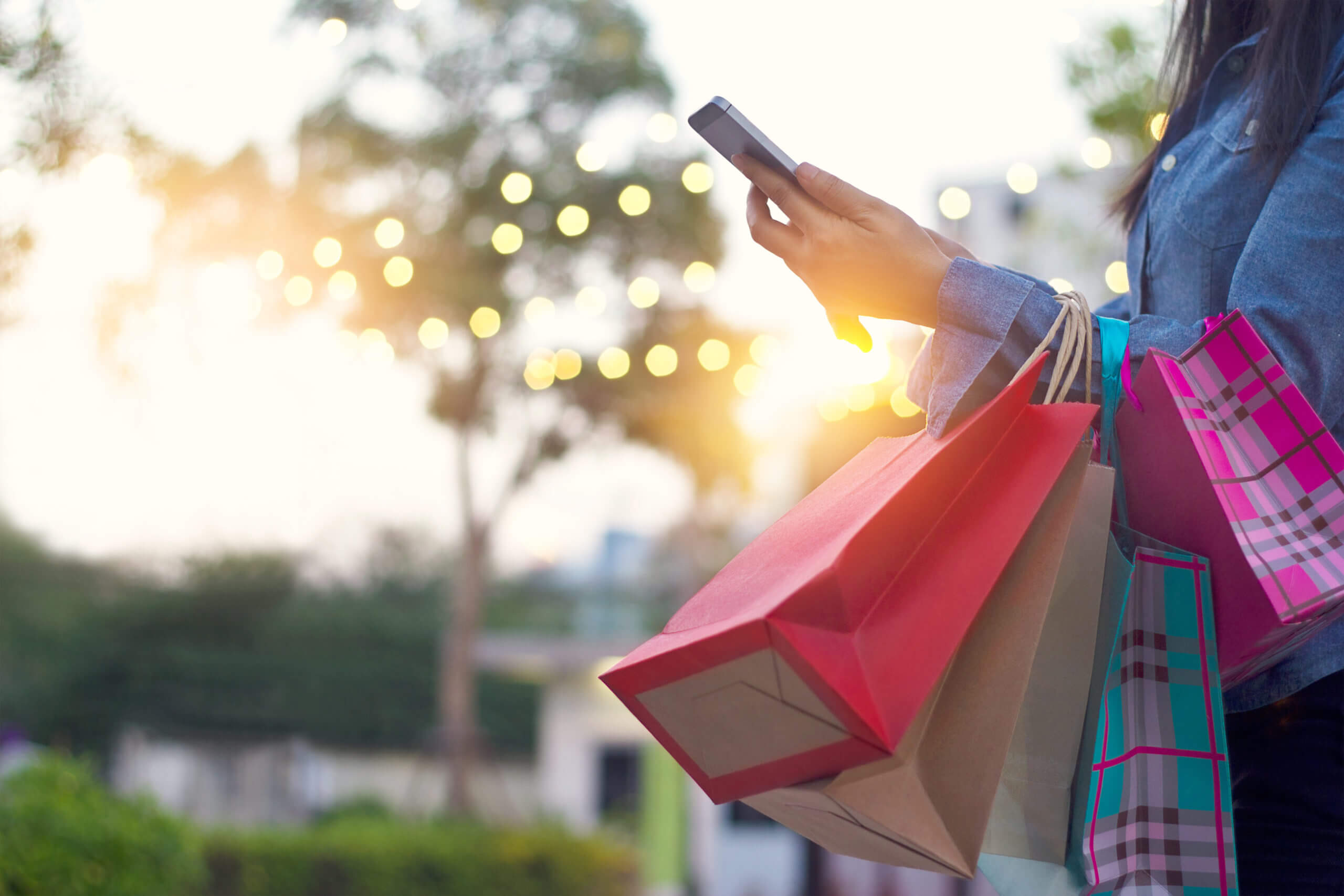 Person wearing a denim shirt holding multiple shopping bags in one arm while using a smartphone outdoors with soft lights and trees blurred in the background at sunset.