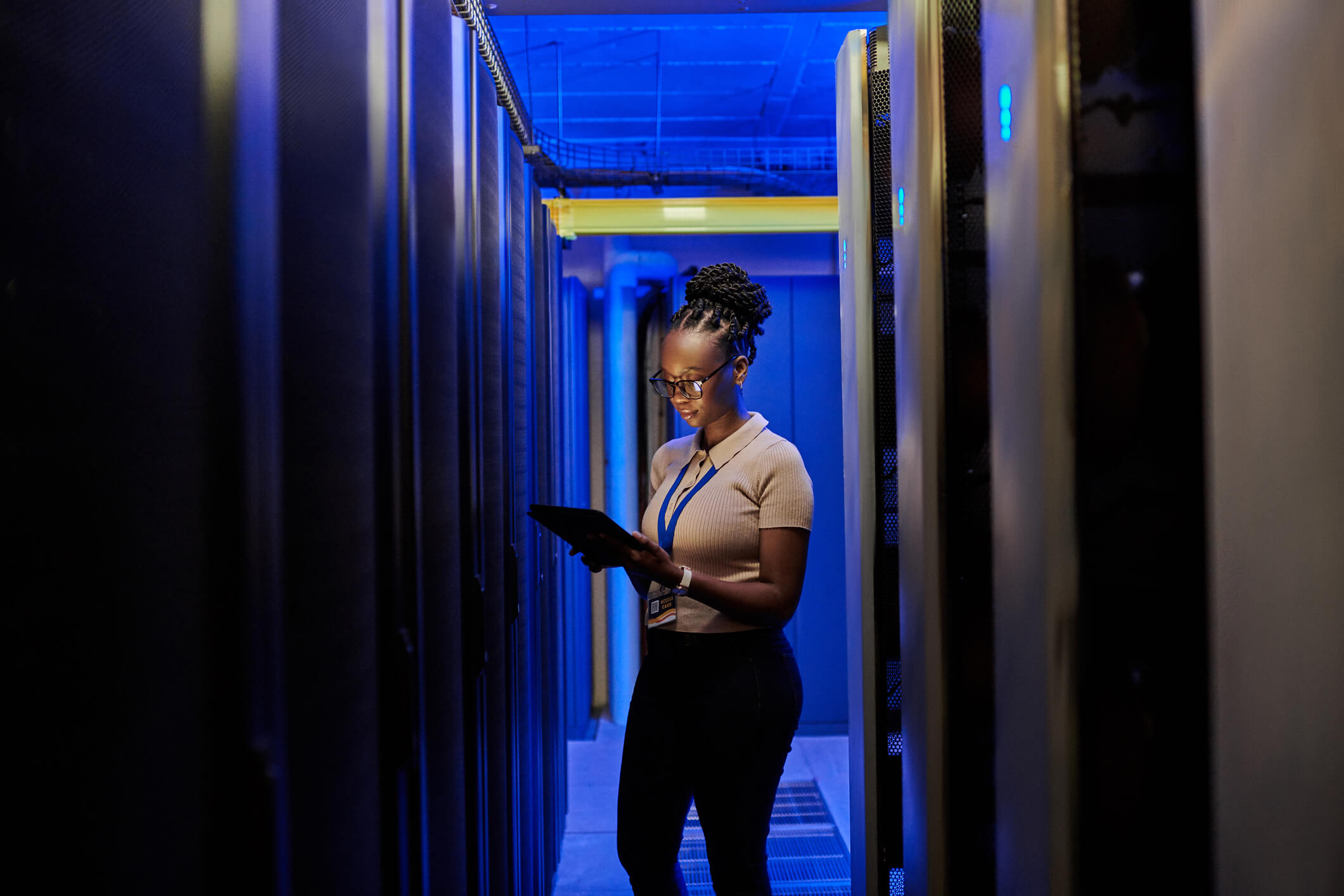 Woman with glasses and braided hair in a beige shirt stands between rows of servers in a data center, holding and looking at a tablet under blue lighting.