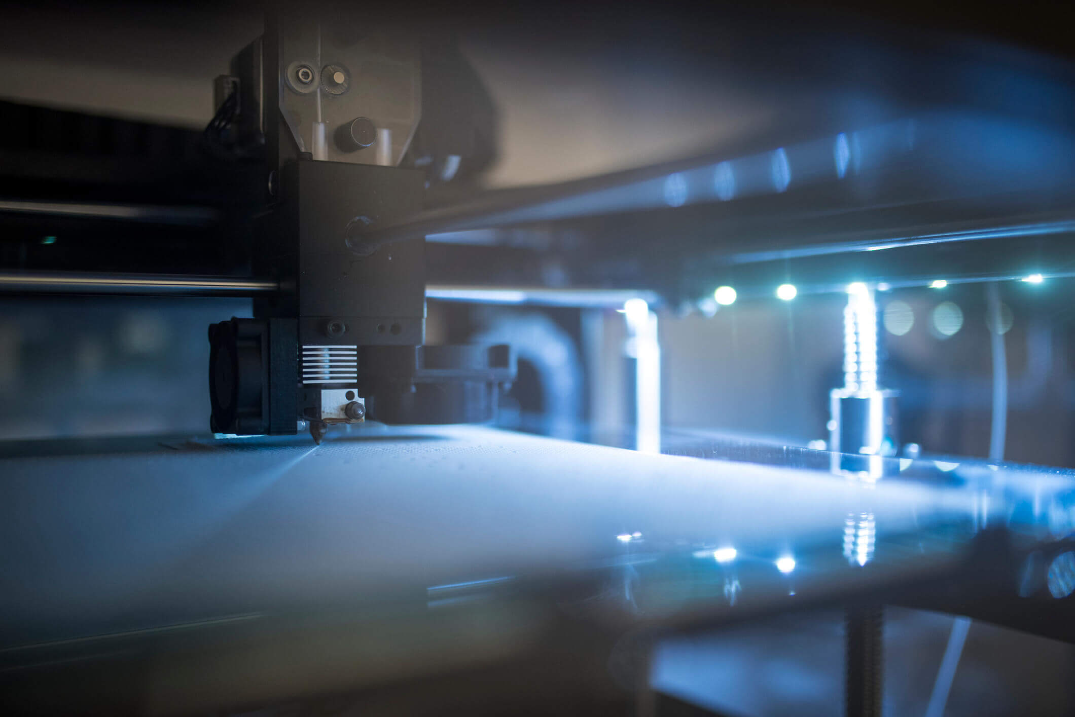 Close-up of a 3D printer nozzle in a dimly lit environment, with blue light illuminating the printing surface and parts of the machine.