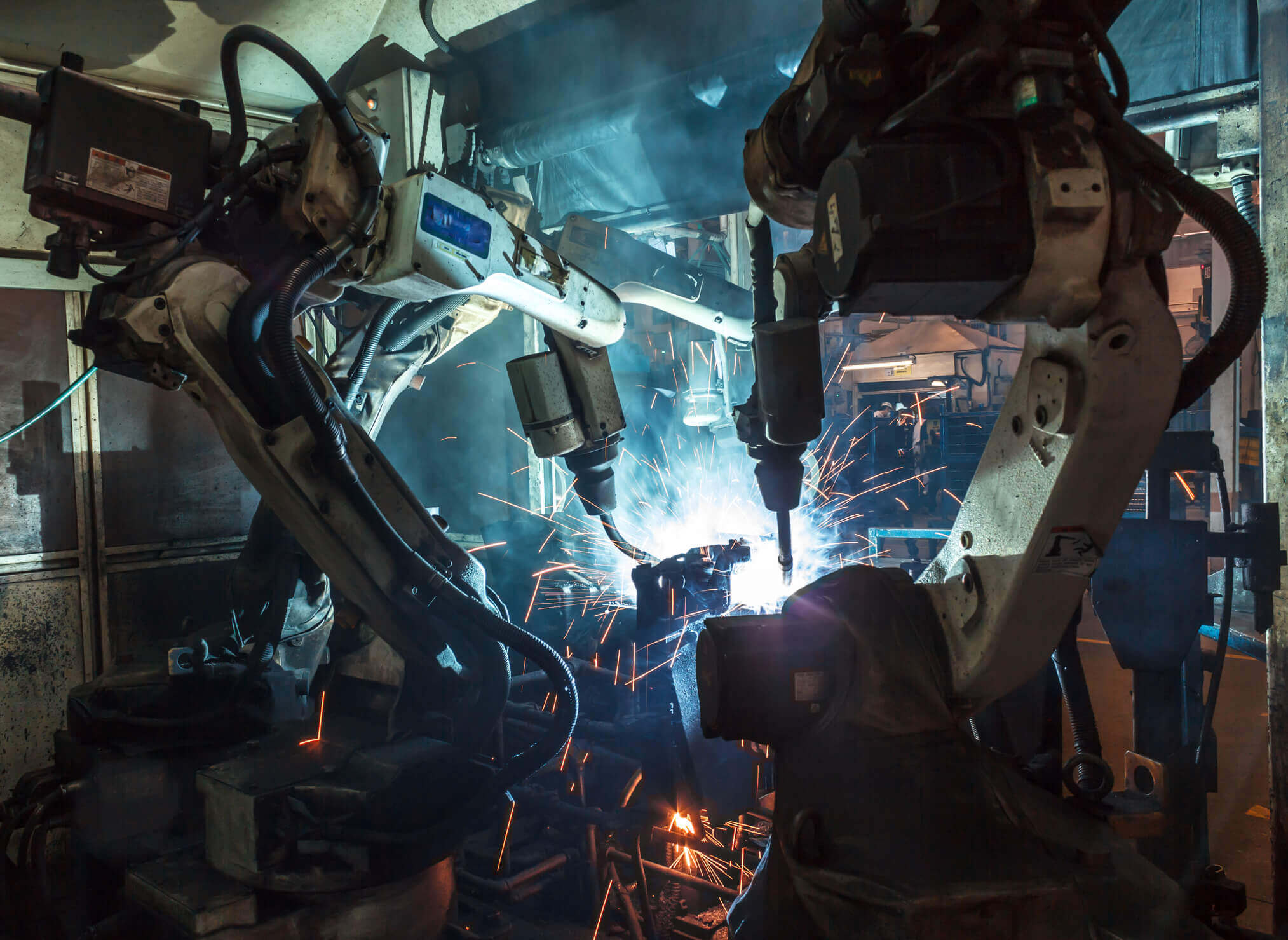 Two industrial robotic arms welding metal parts with bright sparks and smoke in a factory setting.