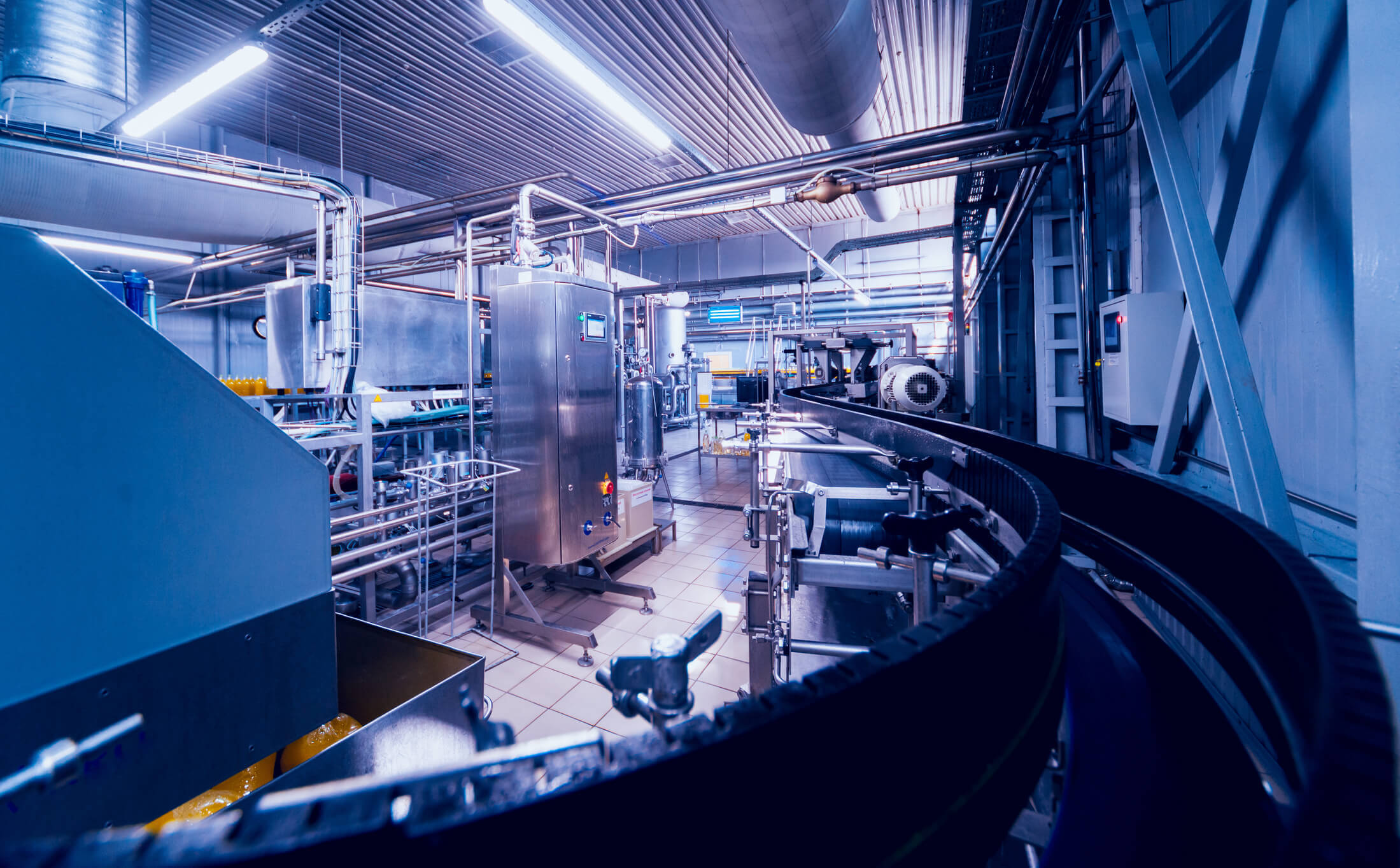 Interior view of a clean, modern industrial manufacturing plant with stainless steel machinery, pipelines, control panels, and a conveyor belt curving through the facility.
