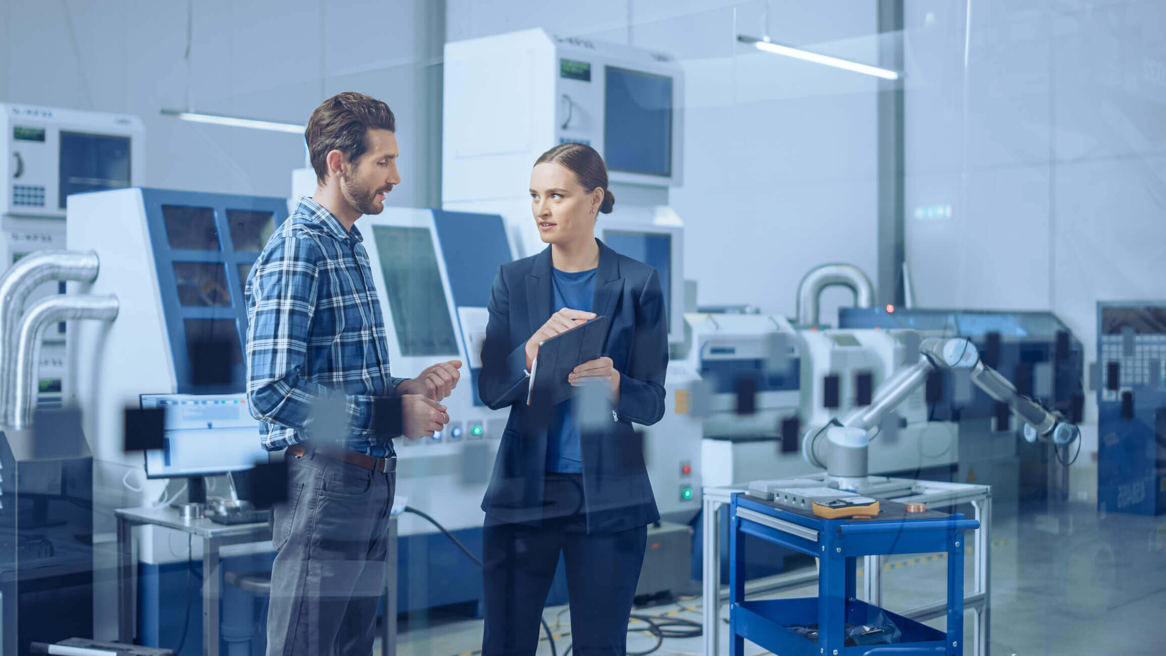Man in a blue plaid shirt and woman in a dark blazer discussing with a tablet in a high-tech industrial manufacturing facility with machinery and robotic arms.