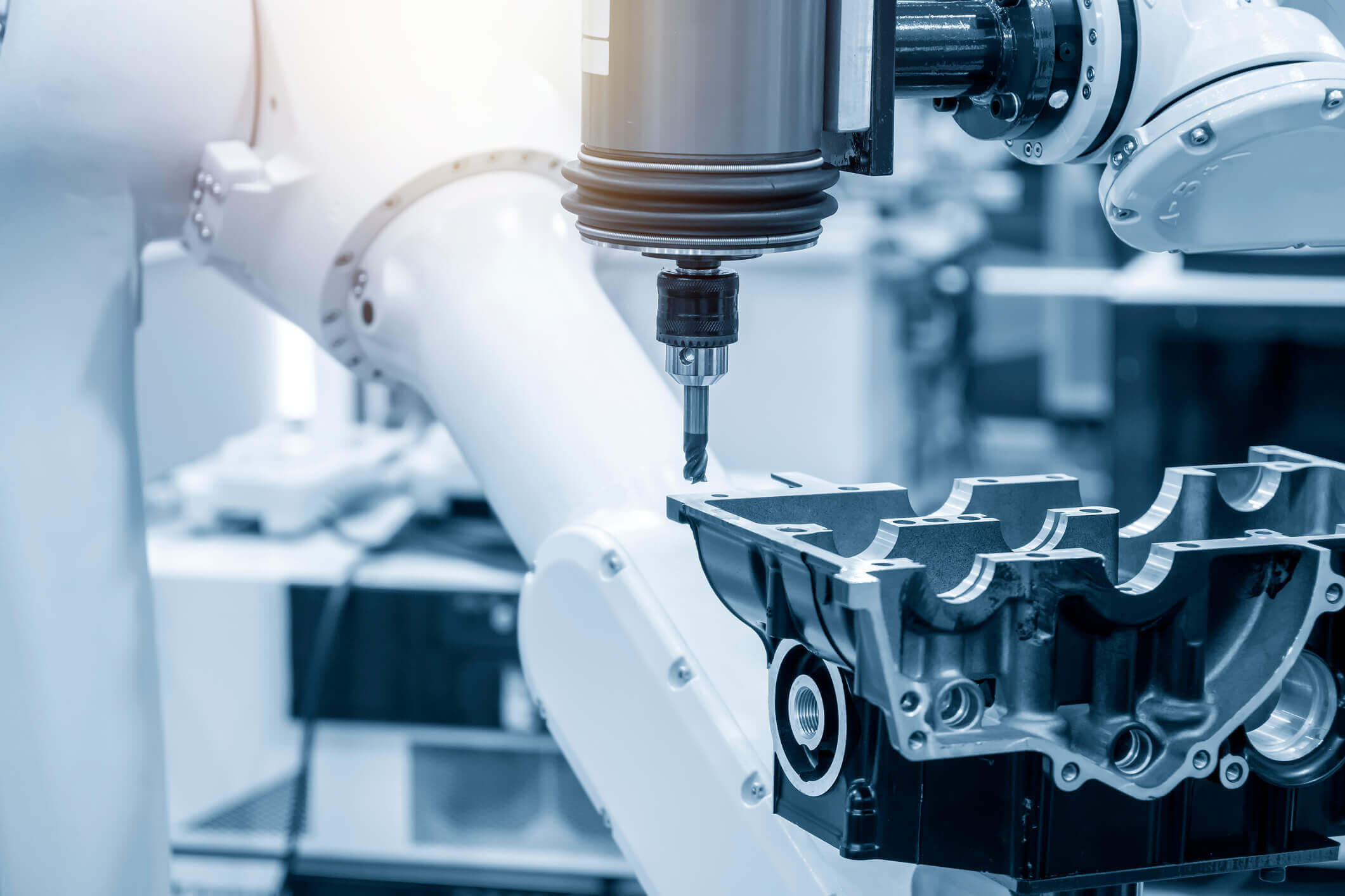 Robotic arm with drill bit operating on metal engine block component inside a manufacturing facility.
