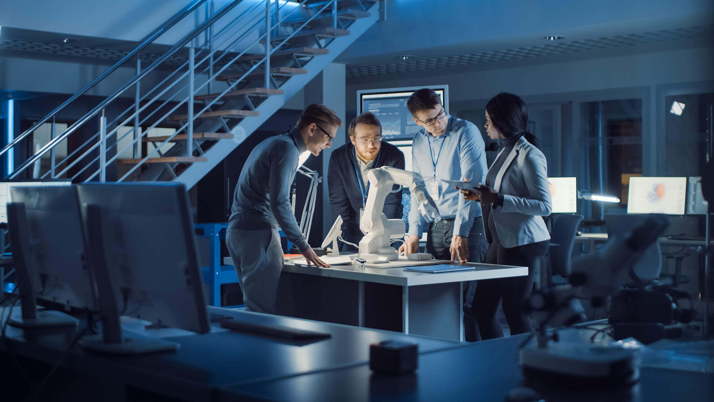 Four engineers in an office study a white robotic arm on a table, surrounded by computer monitors and scientific equipment.