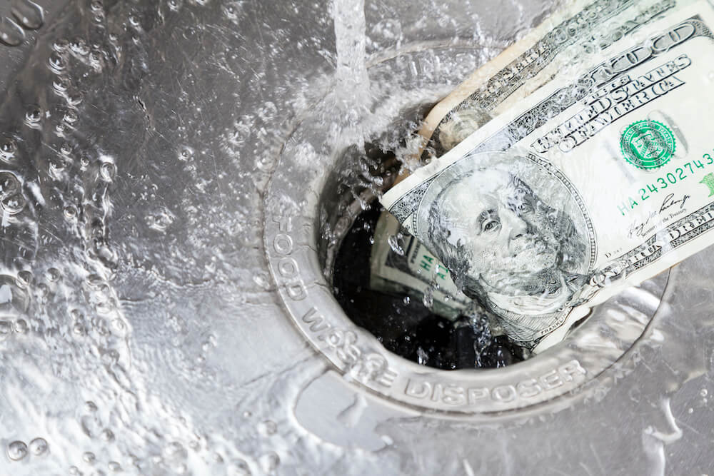 Close-up of a one hundred dollar bill being flushed or dropped into a stainless steel garbage disposal sink drain with water flowing over it.