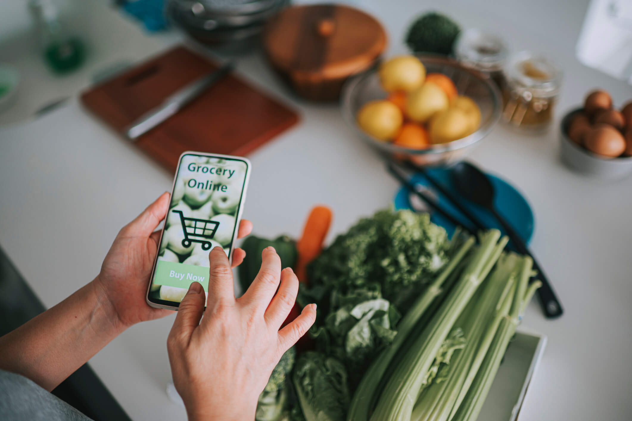 Person using a smartphone with a grocery shopping app showing a shopping cart icon and 'Buy Now' button, with fresh vegetables and eggs on a kitchen counter in the background.