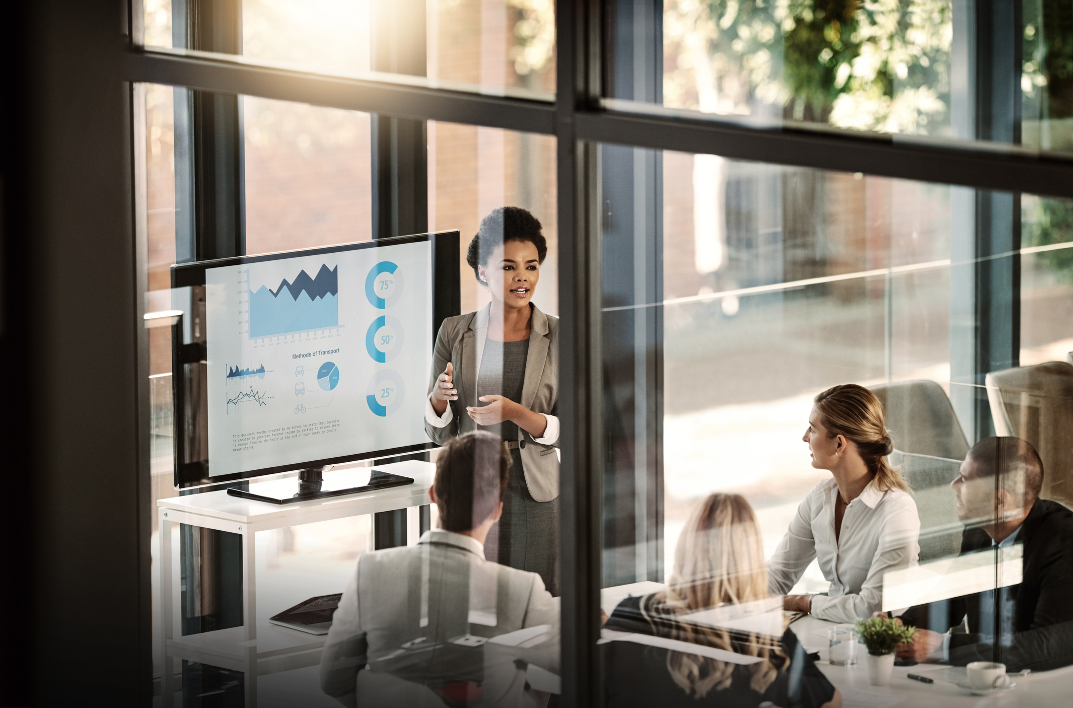 Businesswoman giving a presentation to colleagues in a glass-walled office, standing beside a monitor displaying graphs and charts labeled Methods of Transport.