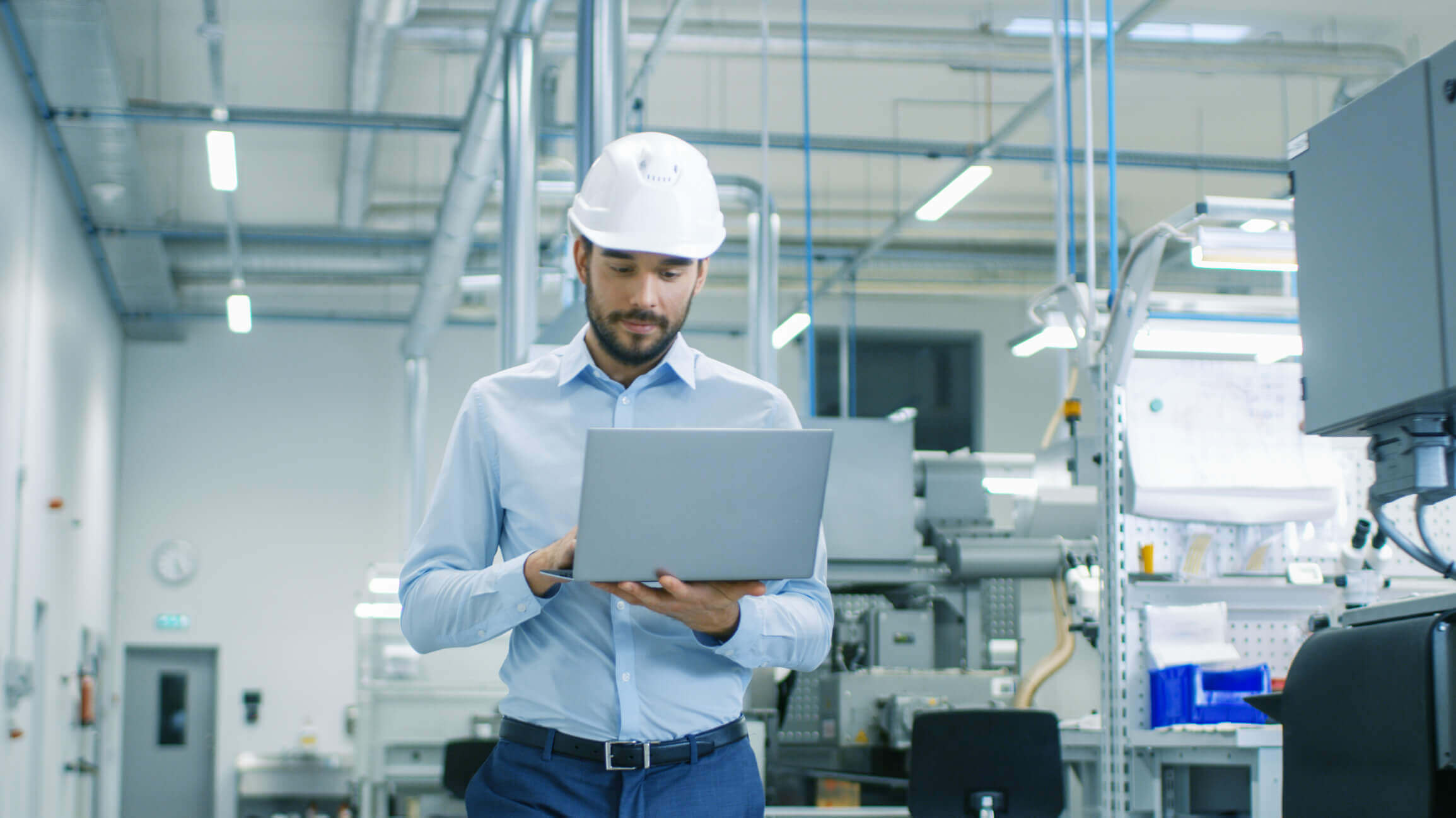 Engineer in a light blue shirt and white safety helmet using a laptop in a modern industrial factory setting with machinery and equipment in the background.
