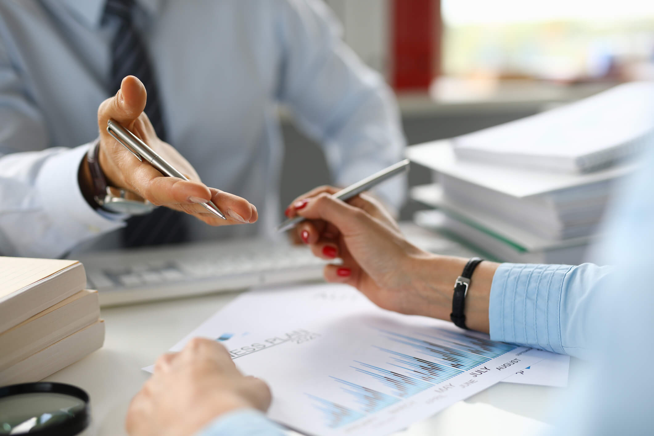 Two business people in a discussion over financial documents with charts and graphs on a desk, one hand holding a pen and gesturing, the other person pointing at the paper with a pen.