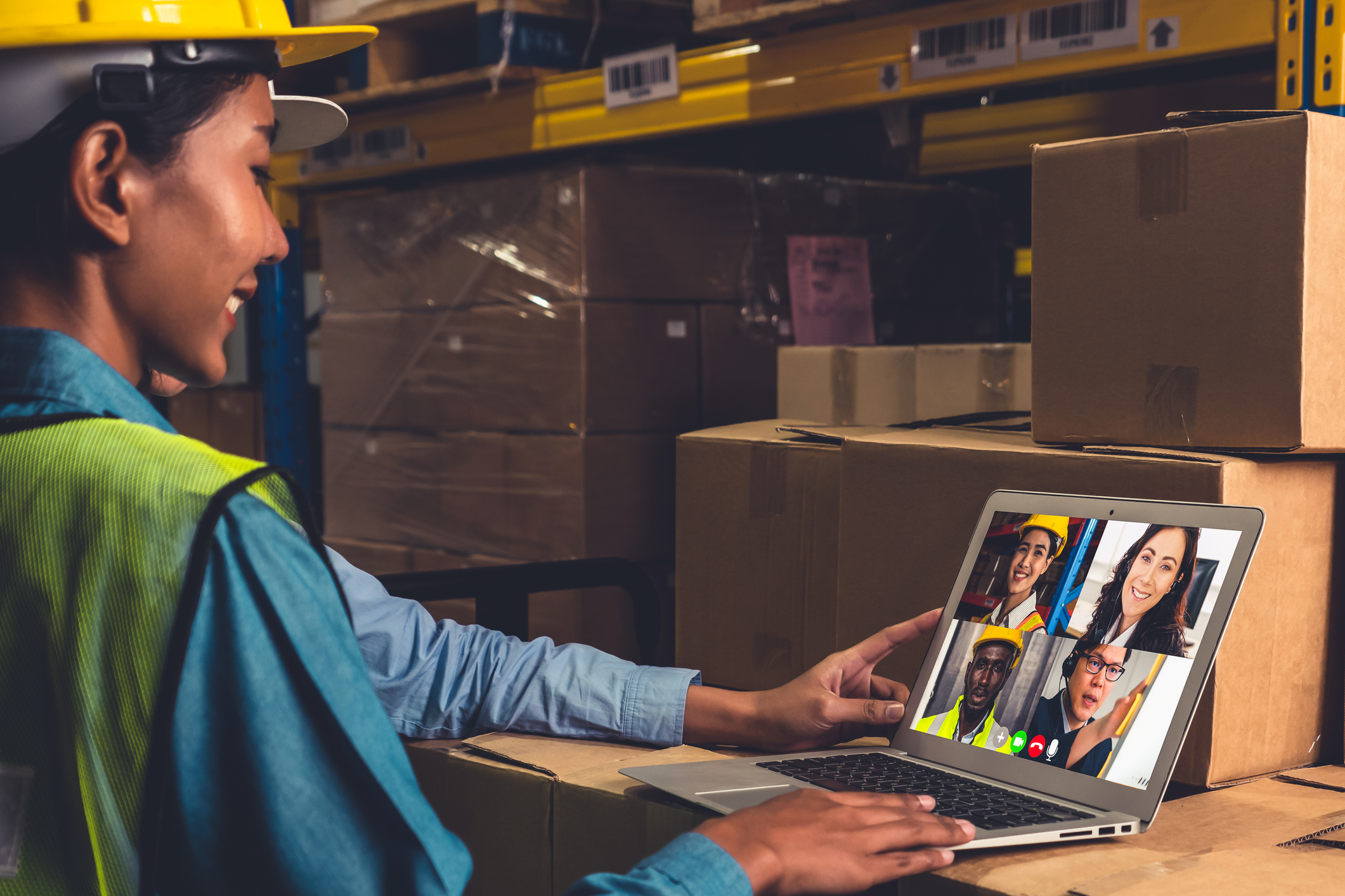 A smiling warehouse worker wearing a yellow safety helmet and reflective vest attends a video conference on a laptop, showing four diverse colleagues on screen, surrounded by stacked cardboard boxes in a storage area.