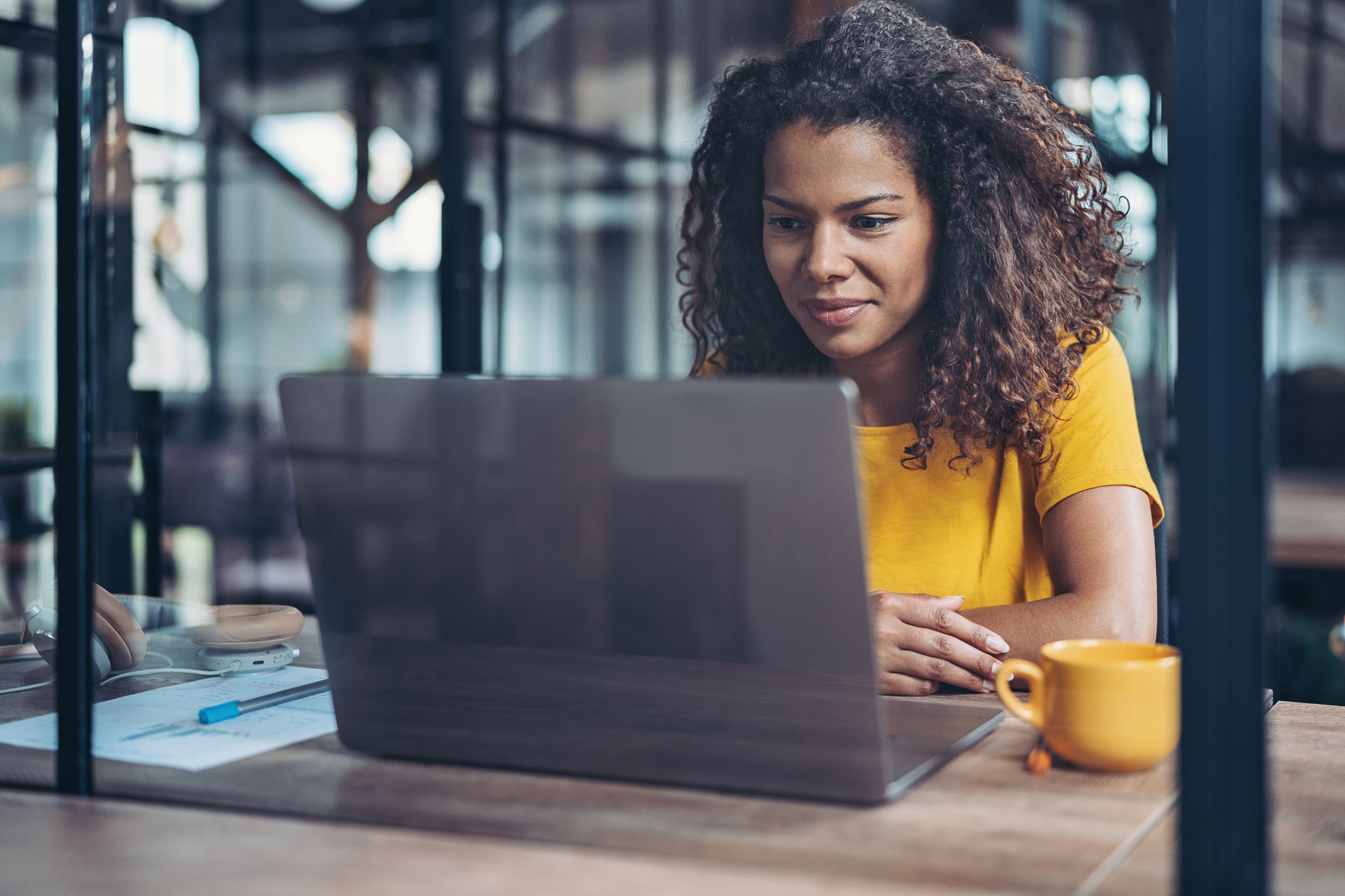 Woman with curly hair wearing a yellow shirt sitting at a table, focused on her laptop screen, with a yellow mug and headphones nearby in a modern indoor setting.