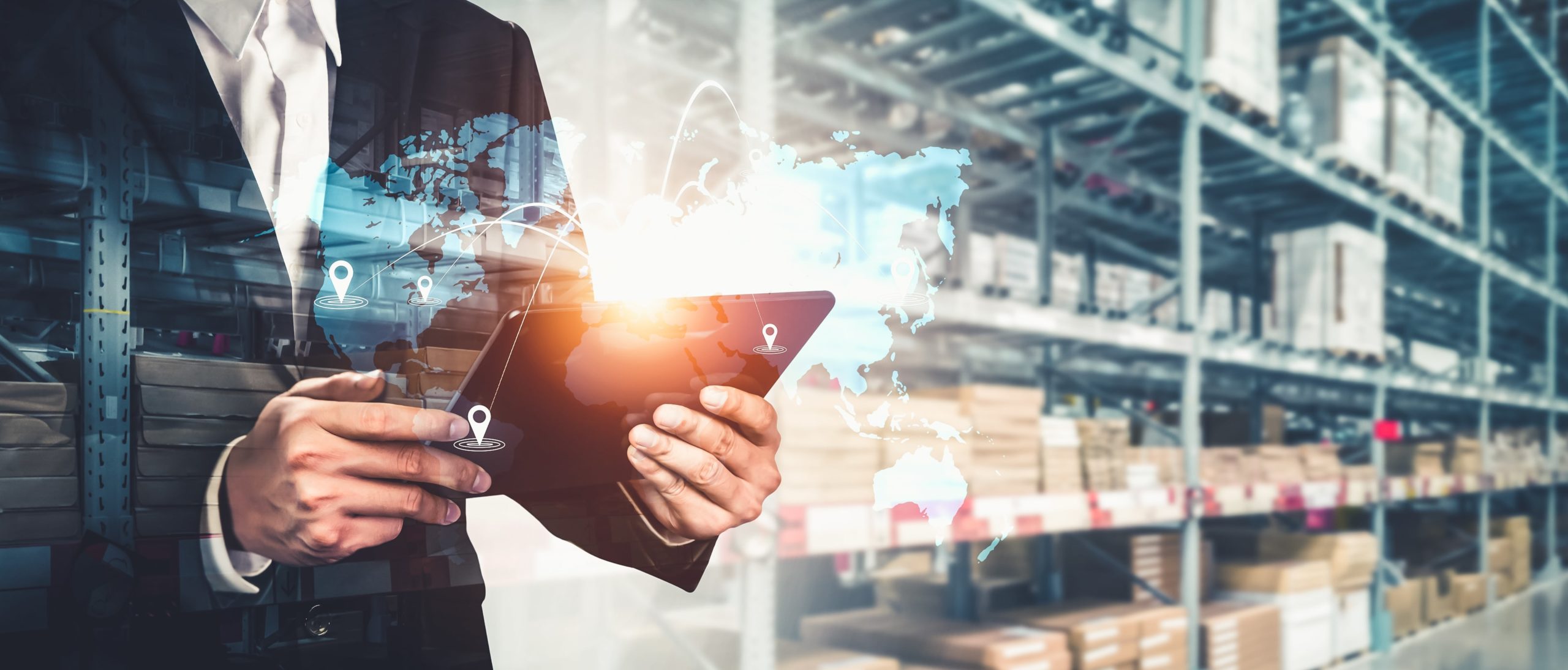 Person in a suit holding a digital tablet with a warehouse background, featuring a transparent overlay of a world map with location pins and connection lines, symbolizing global logistics and supply chain management.