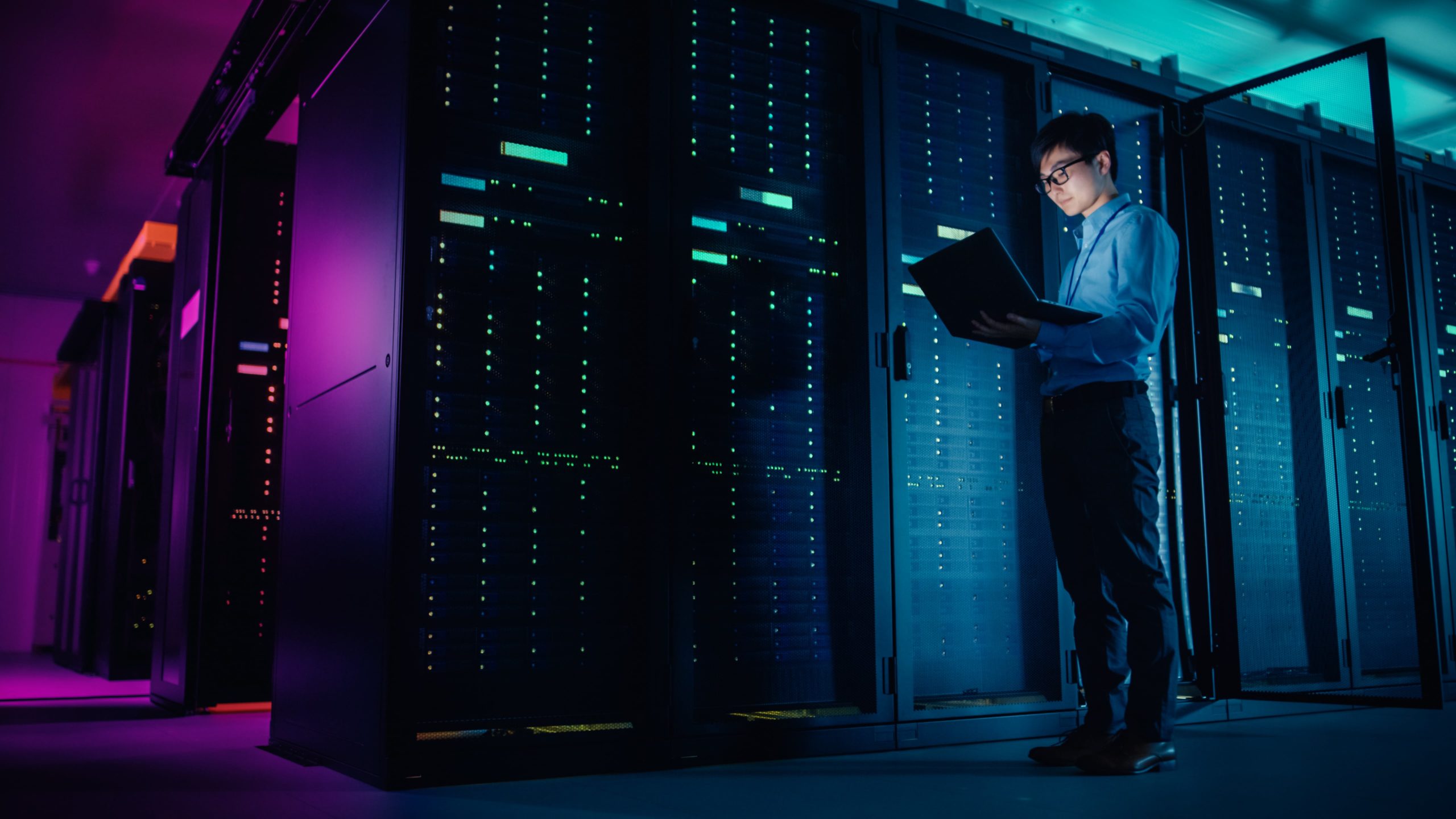 A man in a blue shirt stands in a dimly lit server room holding and looking at a laptop, surrounded by tall racks of servers with illuminated indicator lights.