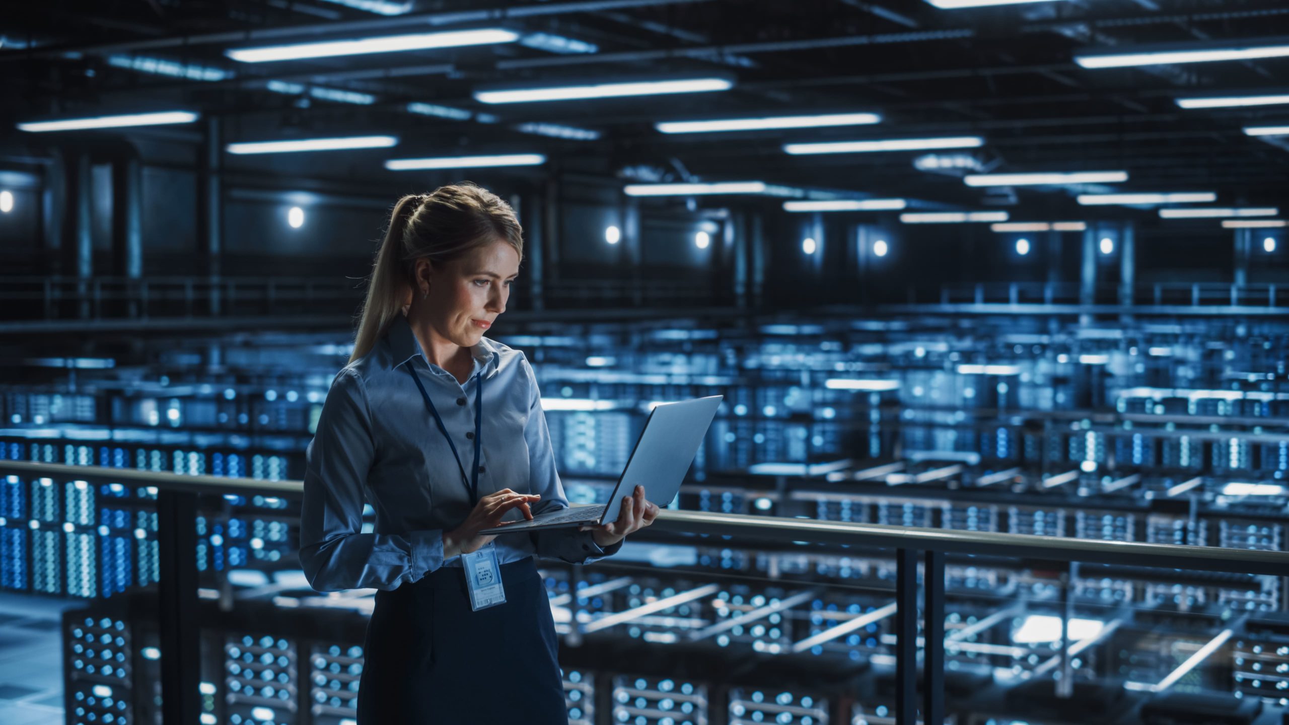 A woman standing inside a large data center, working on a laptop and looking at the screen with focus; the data center has rows of illuminated server racks under bright ceiling lights.