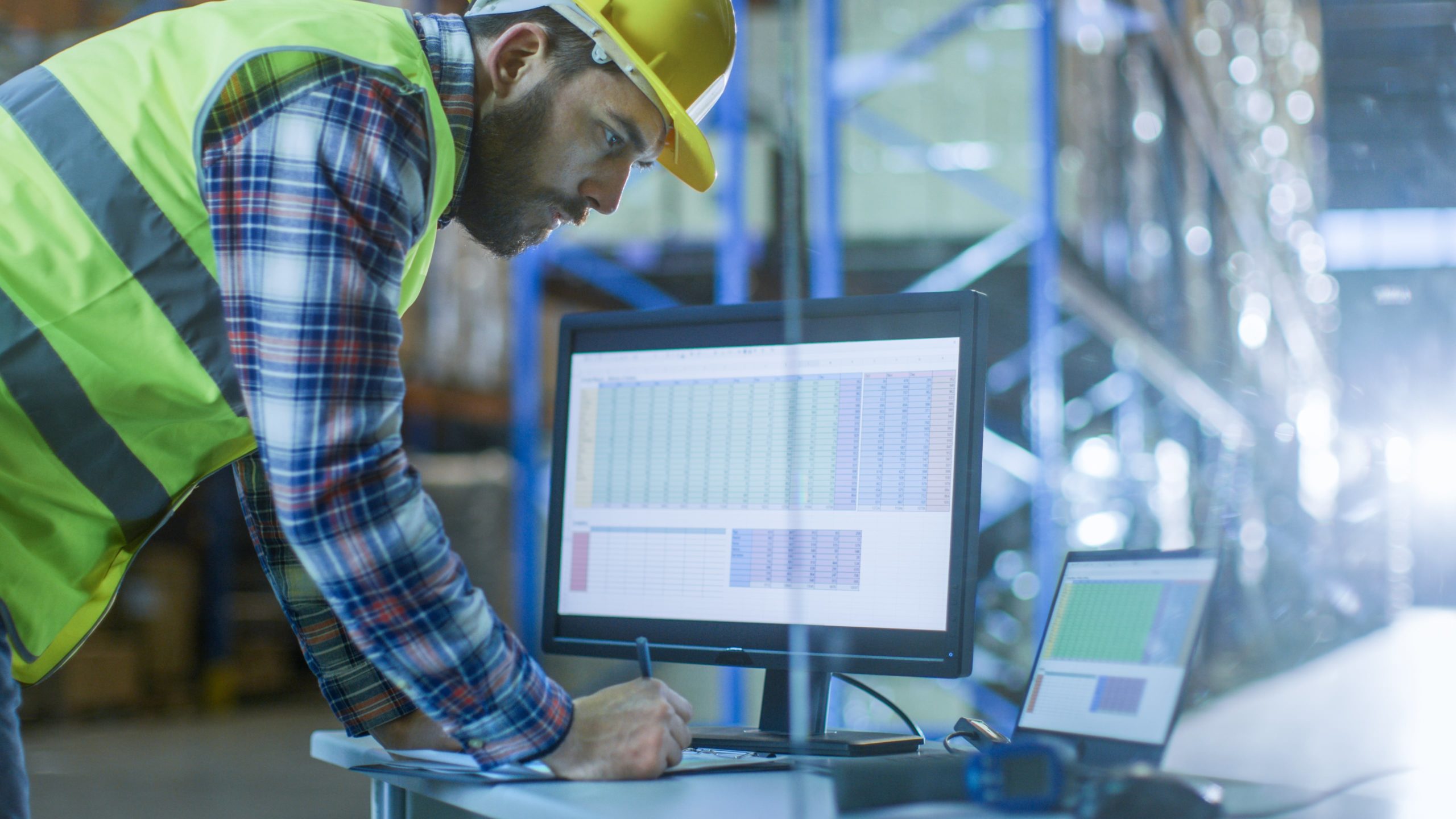 Man wearing a yellow hard hat and neon safety vest working at a desk with a computer monitor and laptop displaying spreadsheets inside a warehouse.