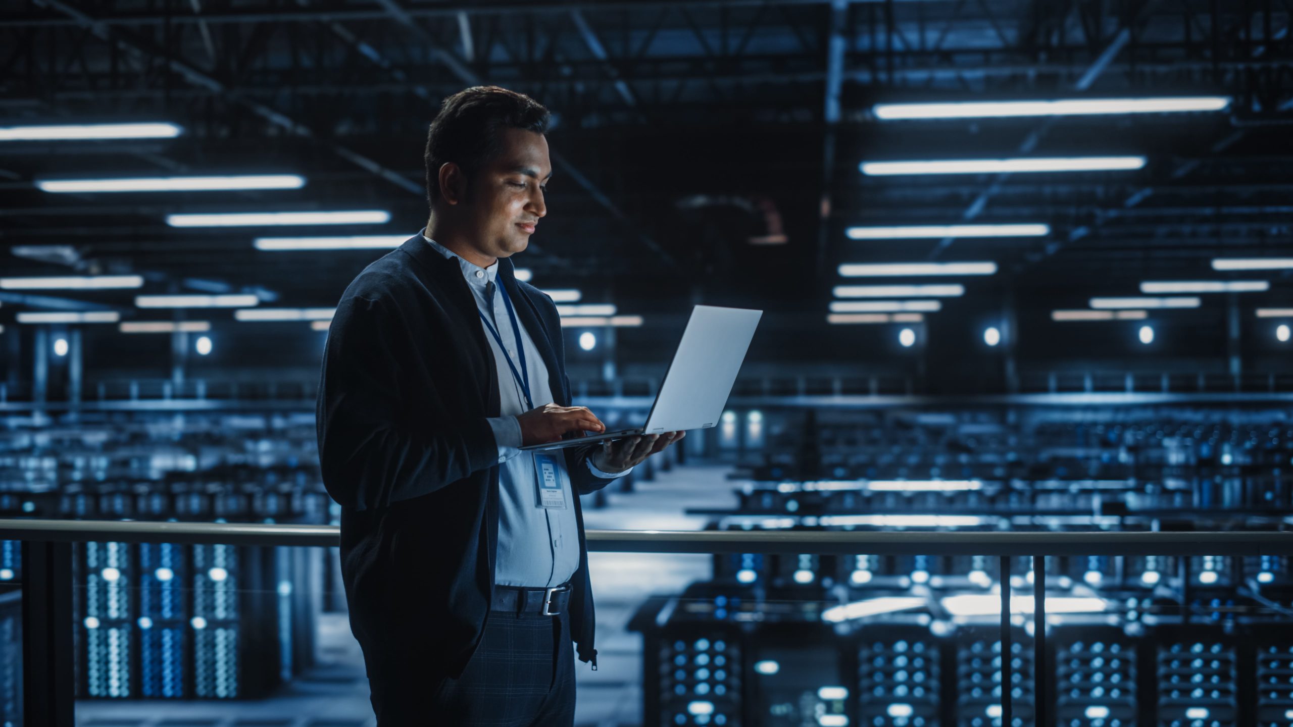 Man wearing a badge stands in a dimly lit data center, working on a laptop with rows of server racks lit in blue lights behind him.