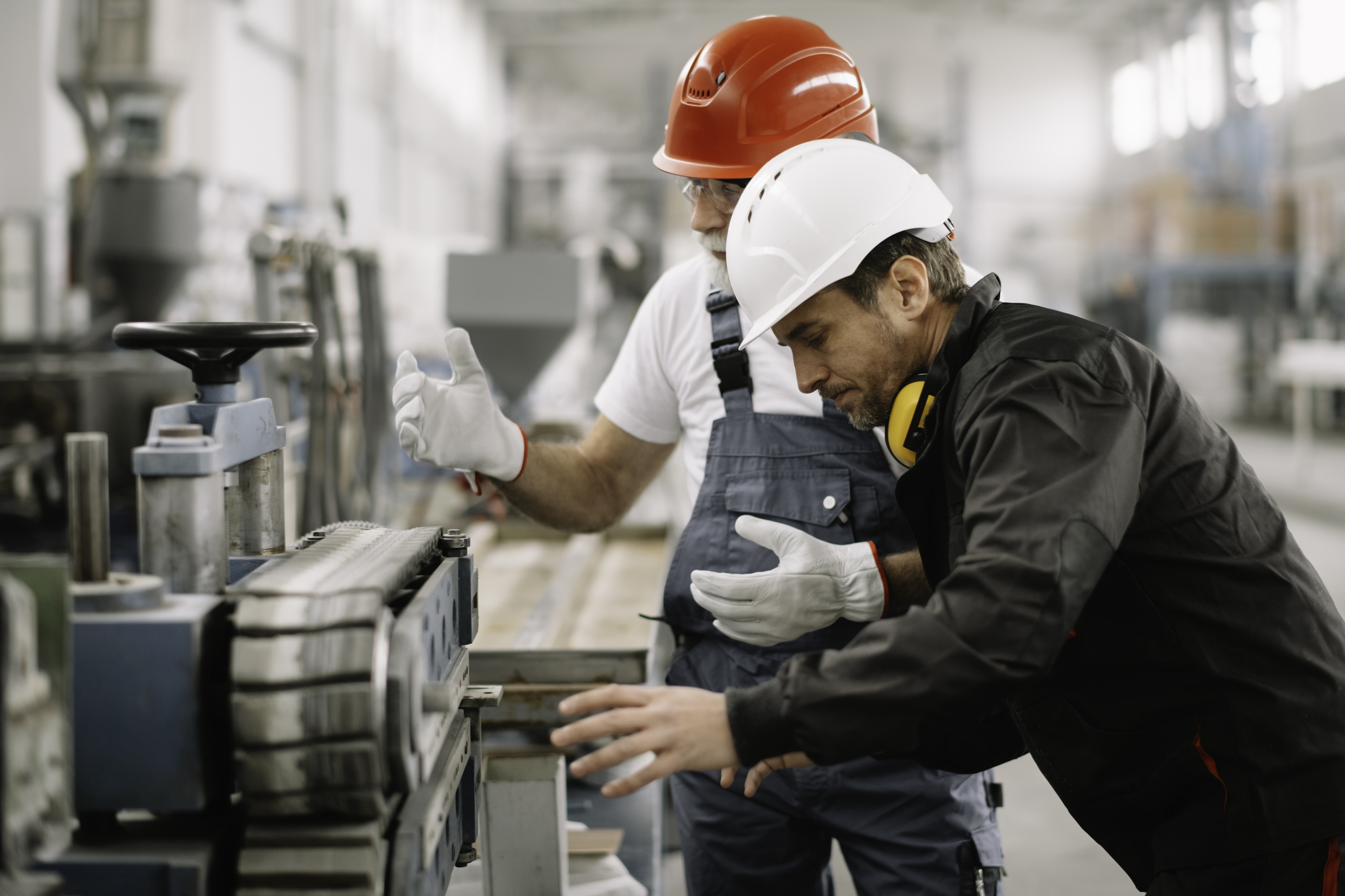 Two factory workers wearing safety helmets and gloves examine and discuss machinery in an industrial setting, with one man gesturing and the other leaning over the equipment.